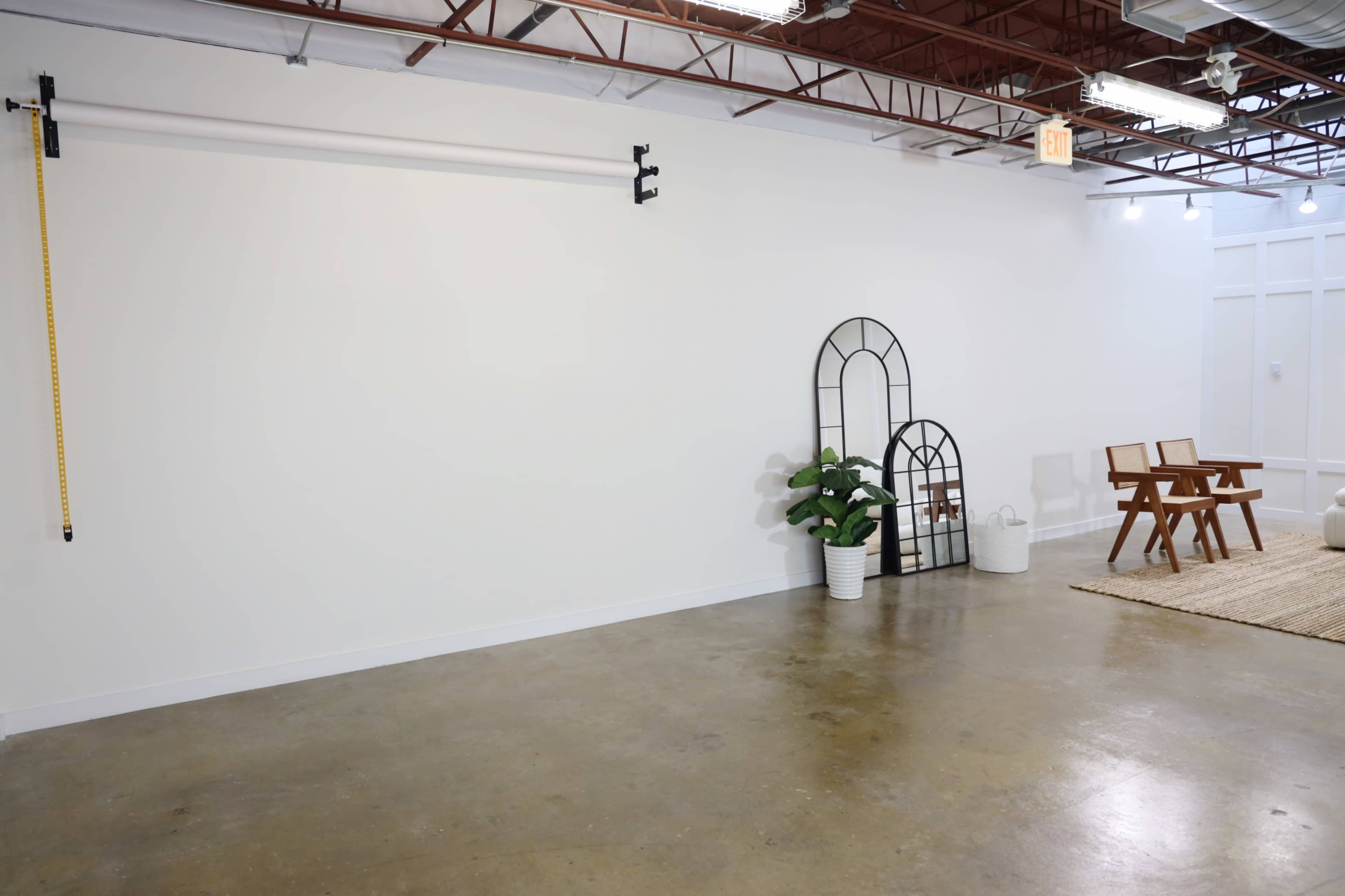 A spacious, empty room with a concrete floor, a few decorative elements including two arched mirrors and a potted plant, and a pair of wooden chairs on a woven rug.