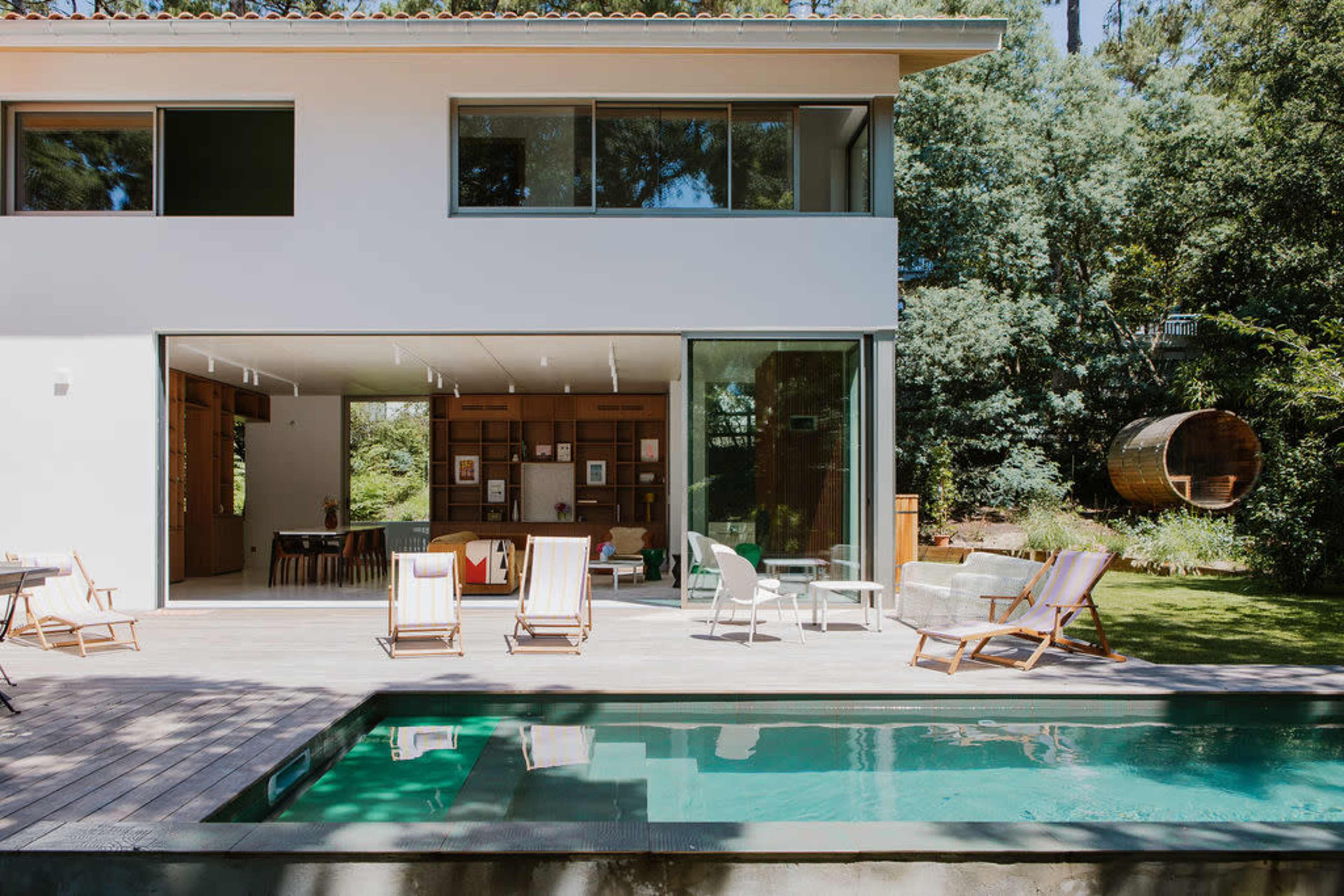 A modern house with large glass doors opens to a patio featuring a swimming pool, lounge chairs, and a grassy yard surrounded by trees.