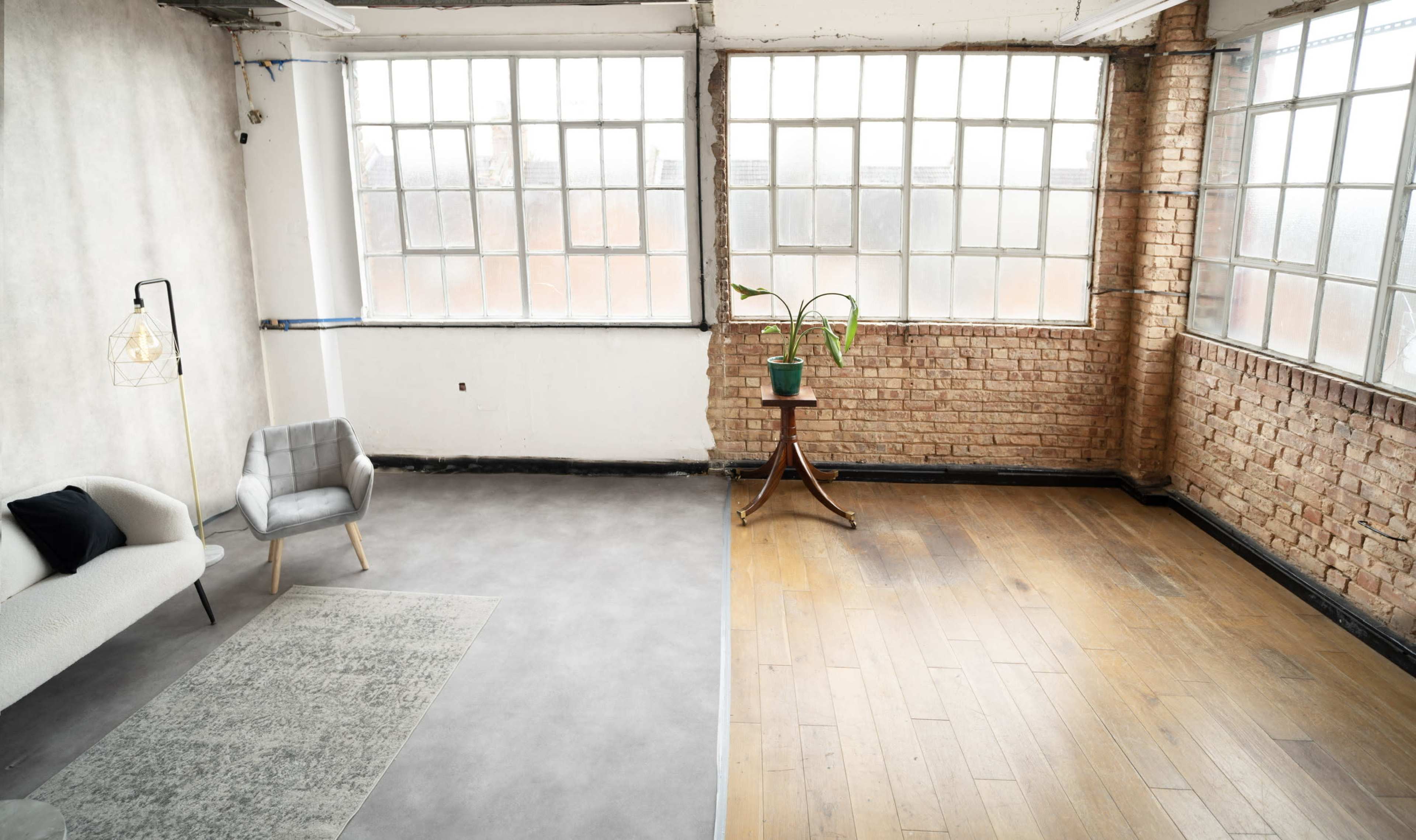 An open space featuring a gray couch and a potted plant next to a wooden table, with large windows illuminating the room.