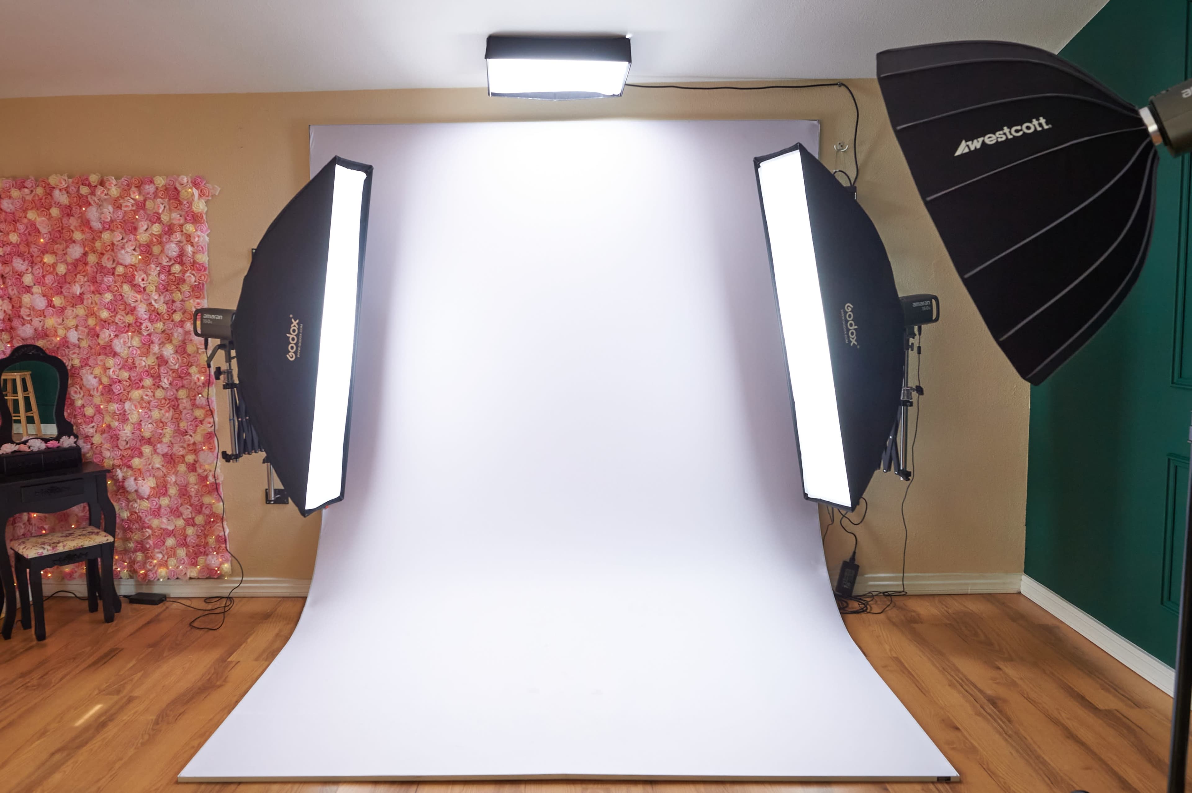 The image shows a photography studio setup featuring a white backdrop, two softbox lights on either side, and a patterned fabric drape in the background.