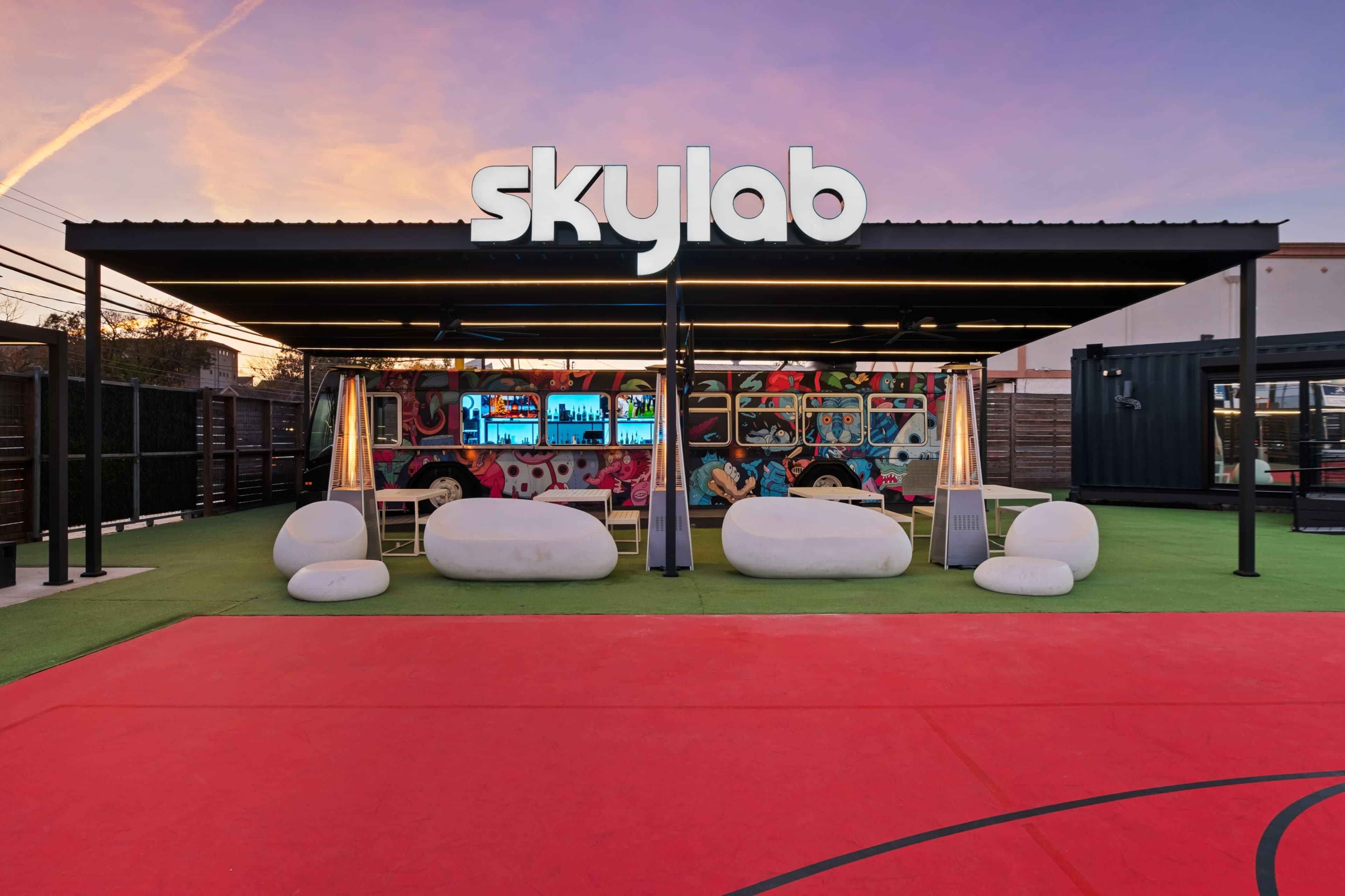 The image shows a colorful outdoor bar area named "Skylab" with modern seating and a vibrant mural in the background, set against a pink and orange sunset sky.