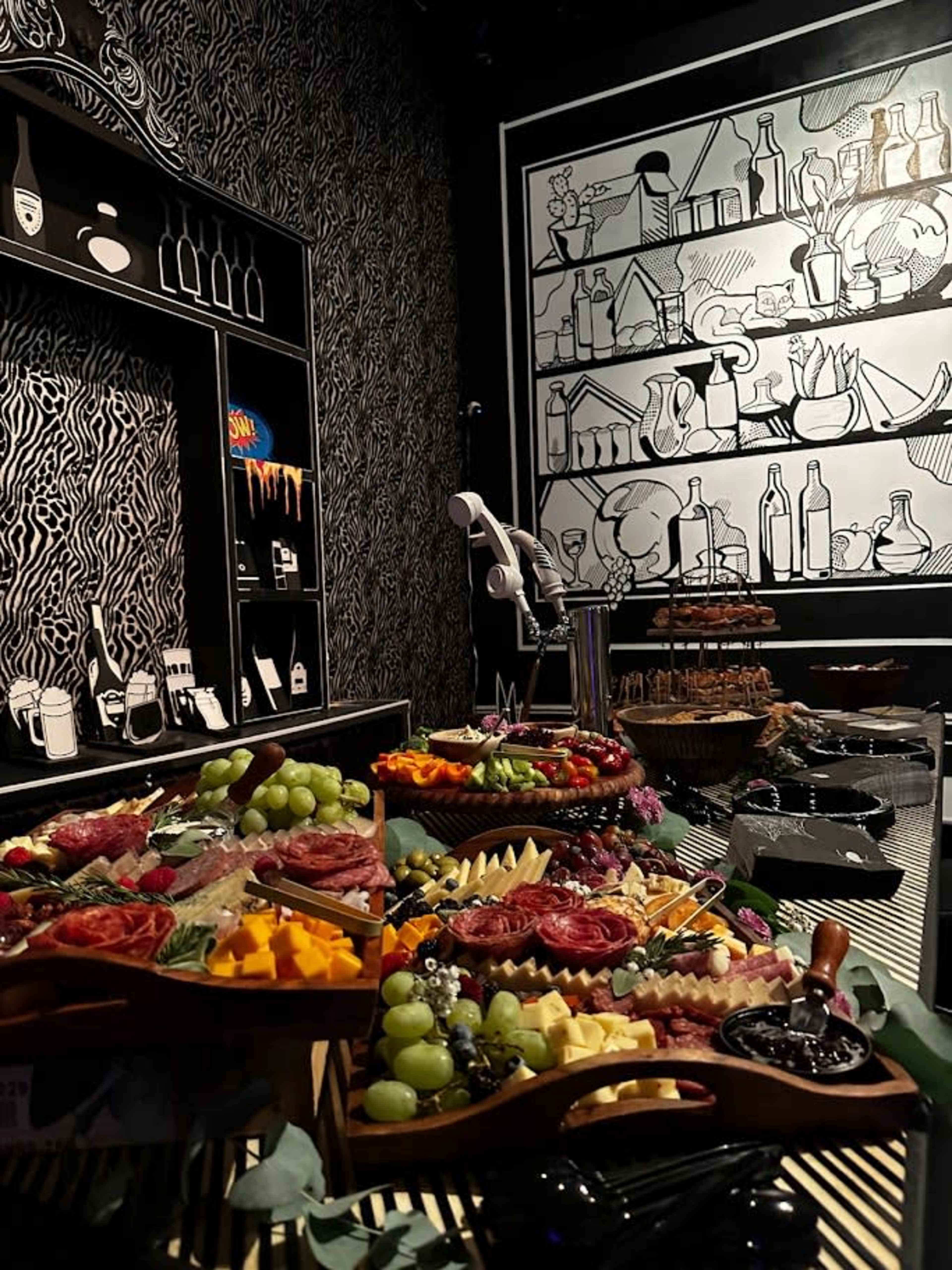 The image shows a vibrant spread of various foods, including fruits, cheeses, and meats, arranged on tables against a backdrop of black-and-white illustrated walls depicting kitchen items.