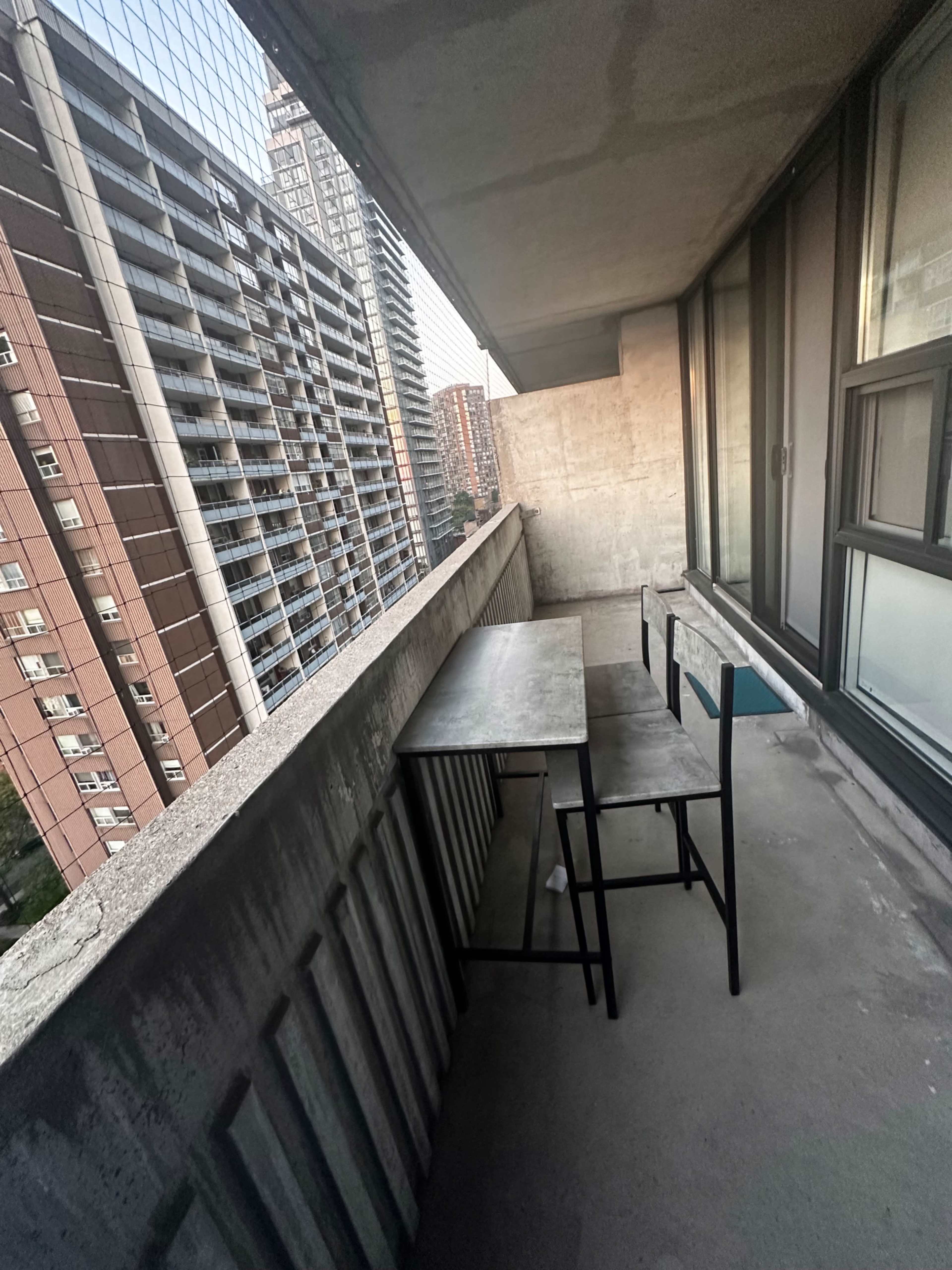 The image shows a balcony with a table and chair overlooking a cityscape of tall buildings.