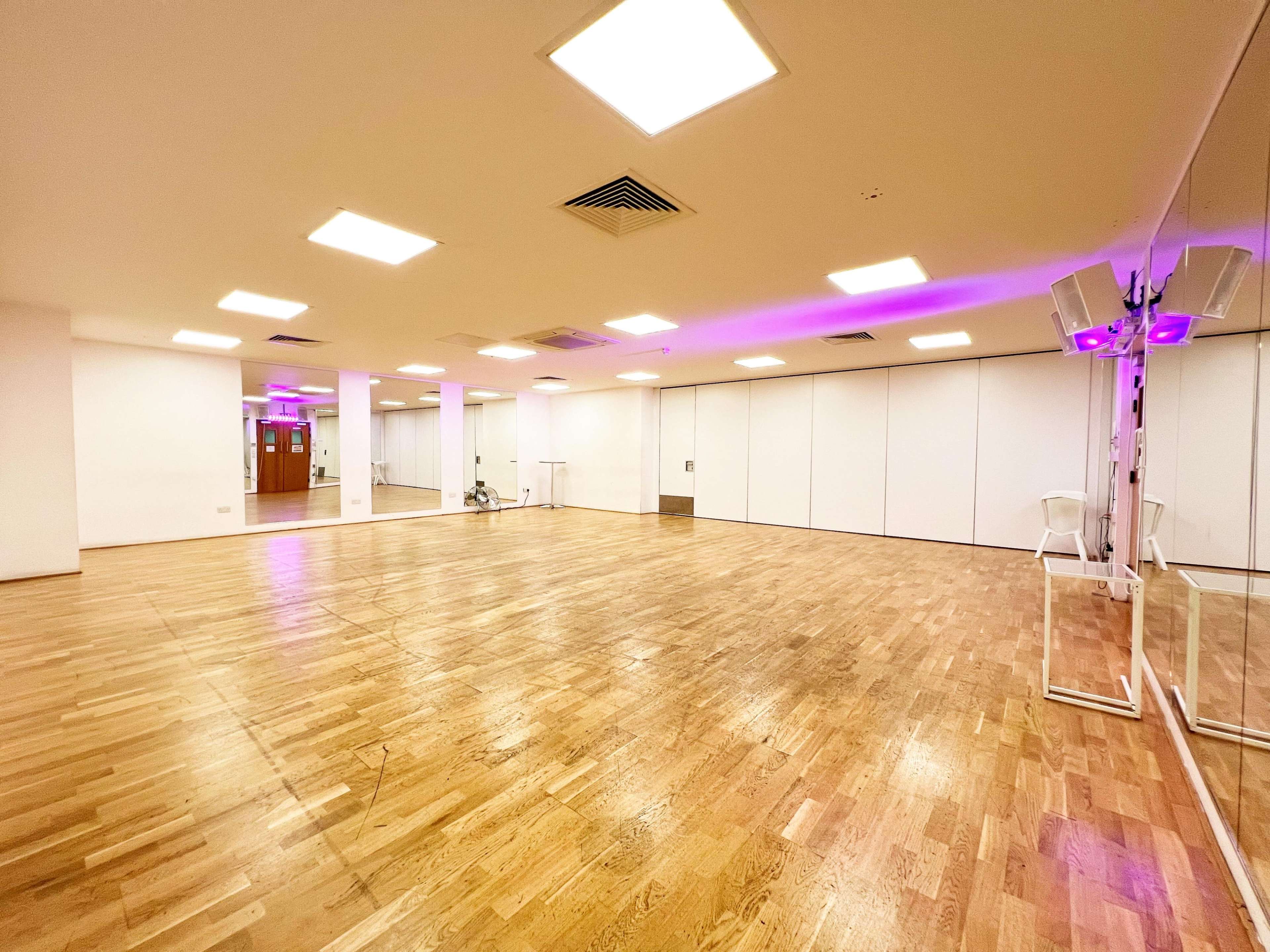 The image shows an empty dance studio with wooden flooring, white walls, and LED lights illuminating the space.