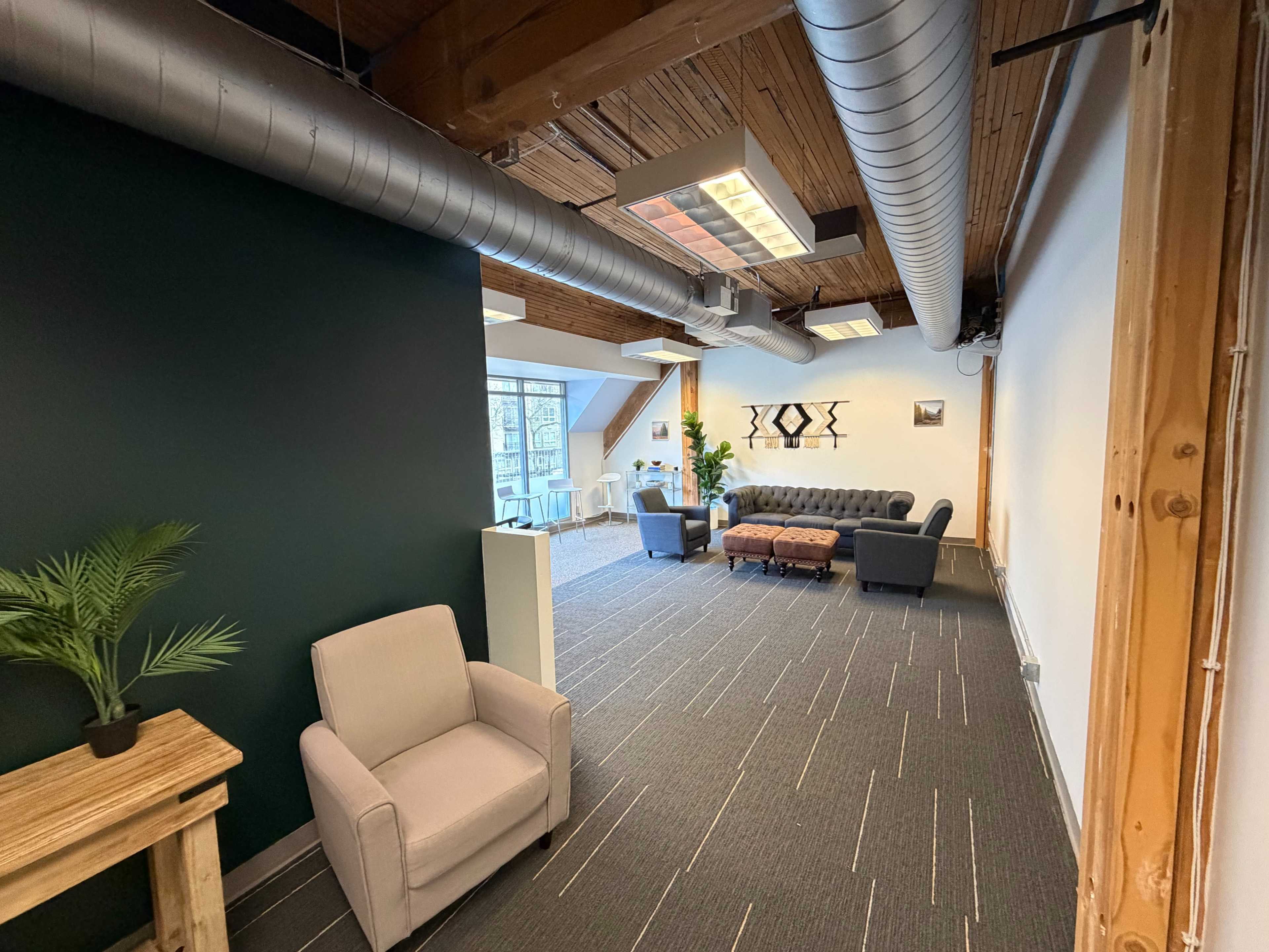 The image shows a modern office space with a seating area that includes a couch and armchair, hardwood beams, and sleek lighting fixtures.