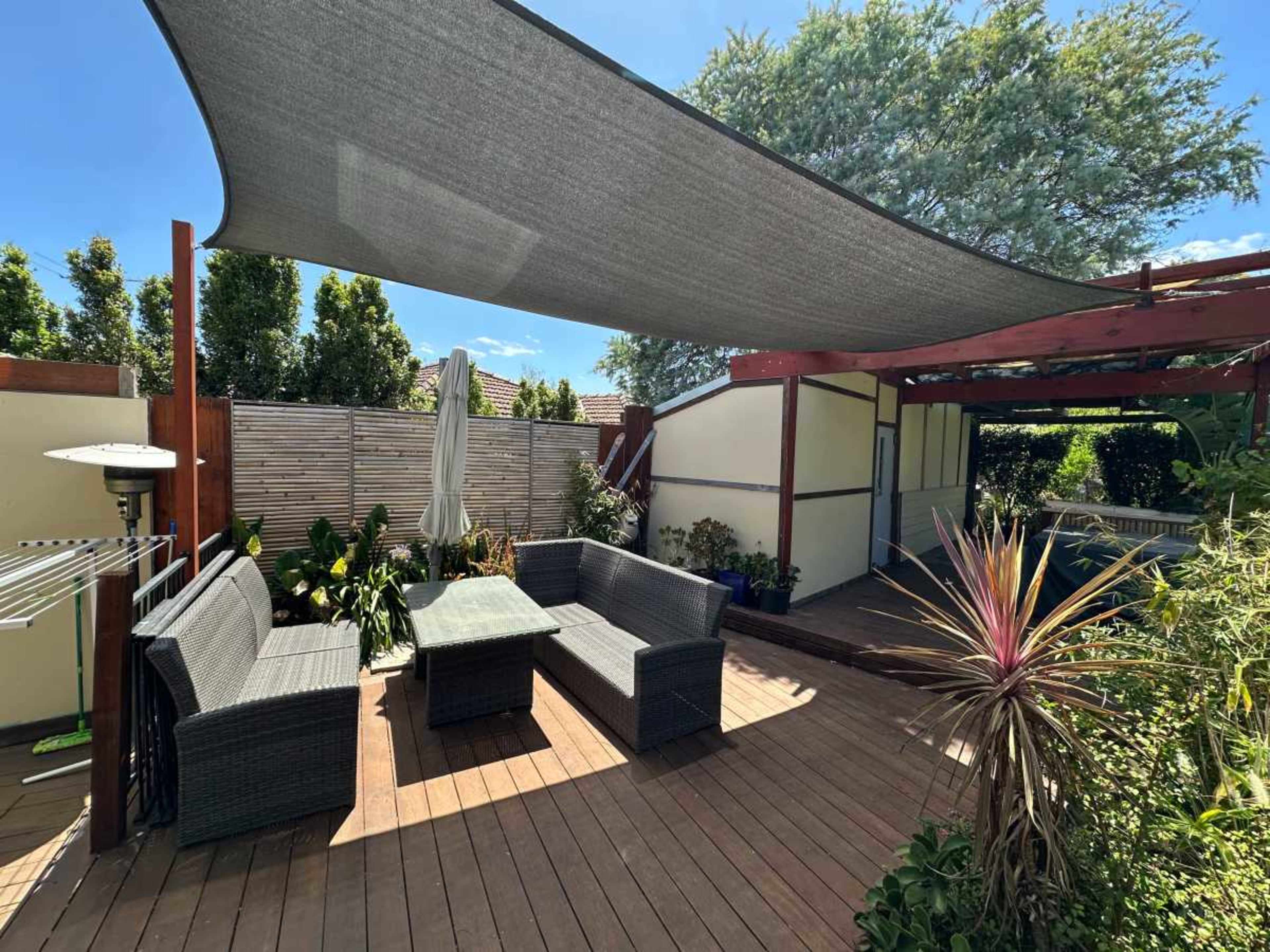 The image depicts a wooden deck patio with a shaded seating area featuring wicker furniture, surrounded by plants and a fence.