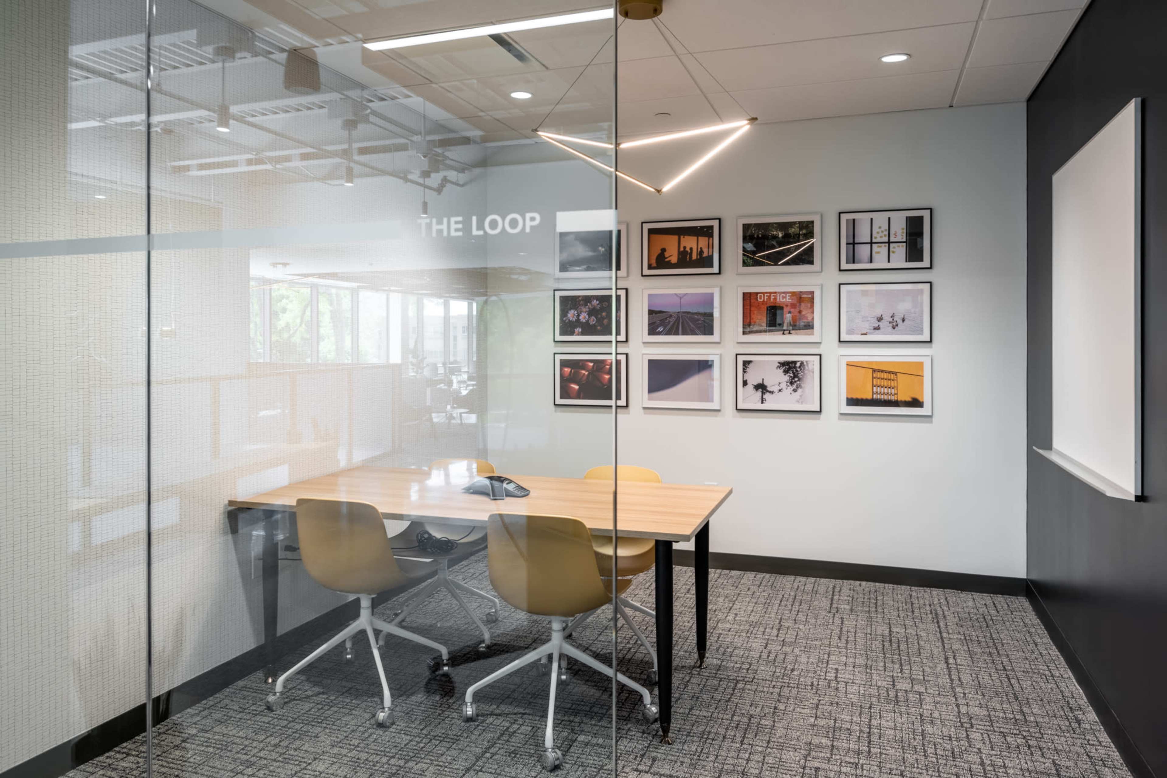 A glass-walled conference room features a wooden table with four chairs and framed photographs on the wall.