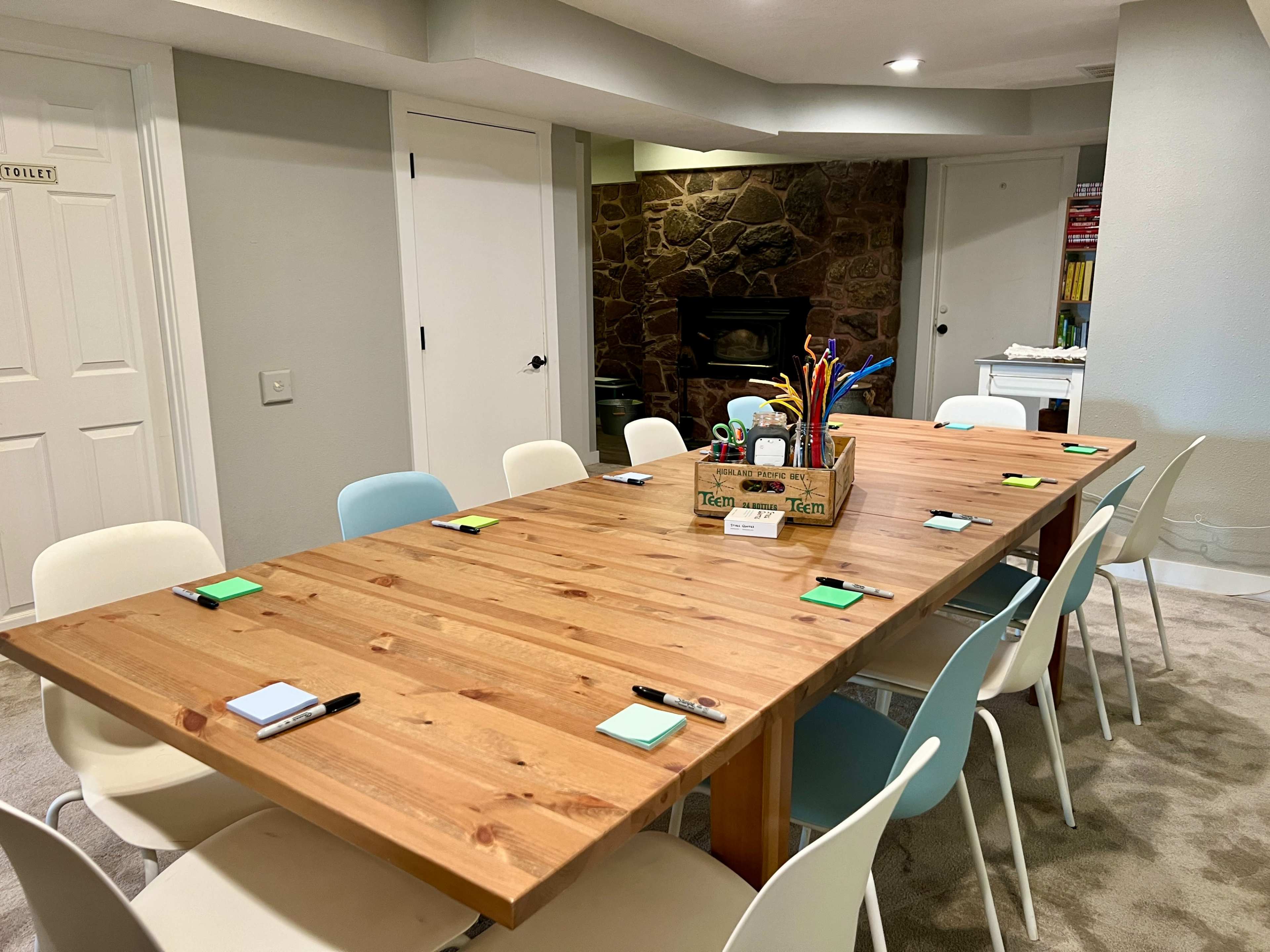 A conference room features a large wooden table surrounded by chairs, with a variety of stationery on the surface and a stone fireplace in the background.