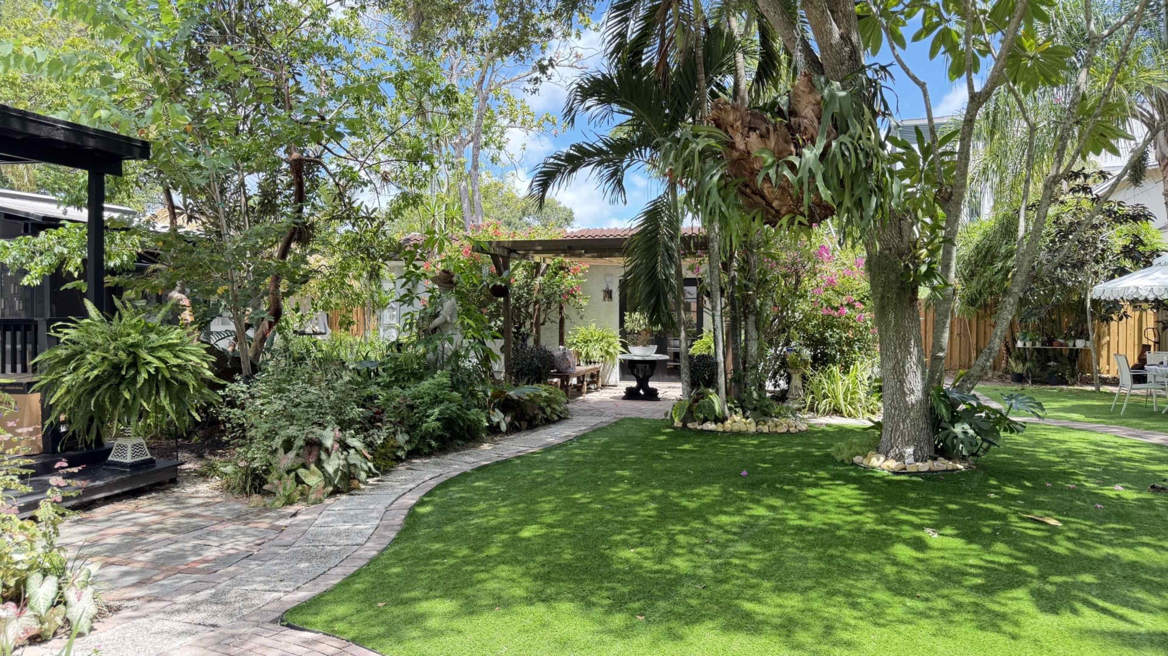 The image shows a landscaped garden with a winding pathway bordered by various plants and trees, leading to a gazebo or seating area in the background.