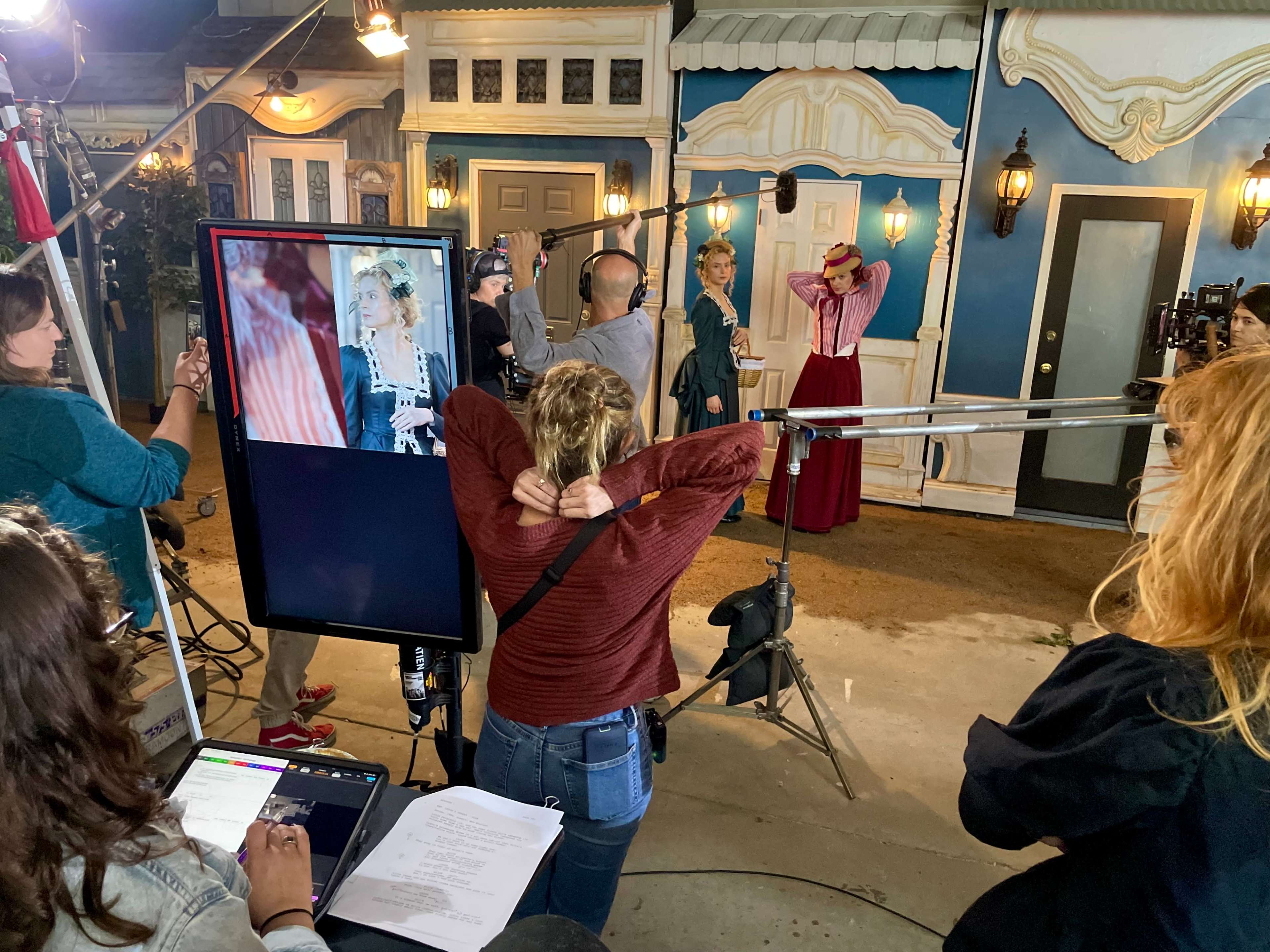 A film crew is set up on a stage with period-style decor, focusing on a woman in historical attire as she poses in front of a monitor.