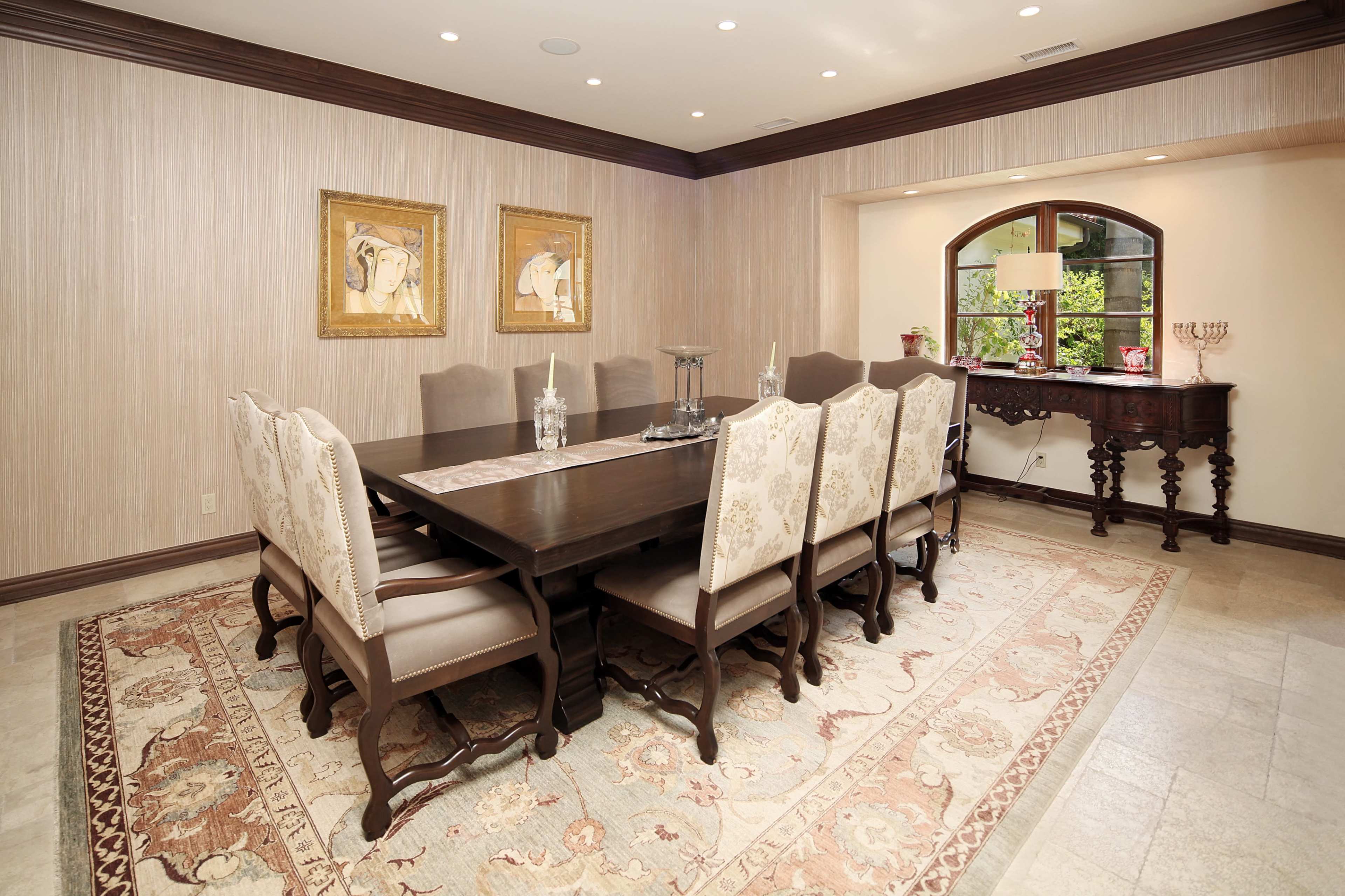 A dining room features a long wooden table surrounded by eight upholstered chairs, with ornate artwork on the walls and a side console table beneath a window.