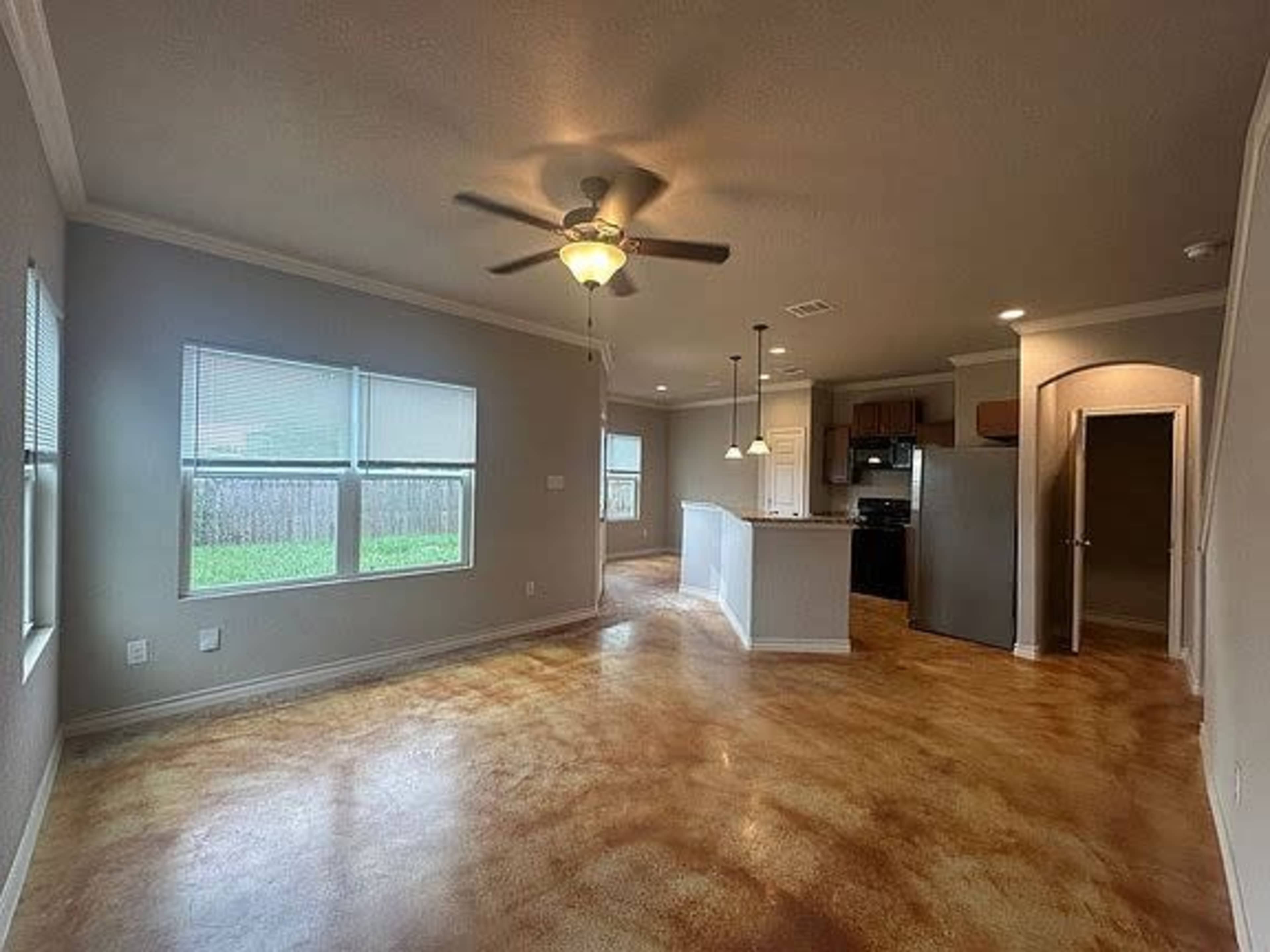 The image shows a spacious living area with a ceiling fan, two windows, and an open layout connecting to a kitchen area featuring a bar and appliances.