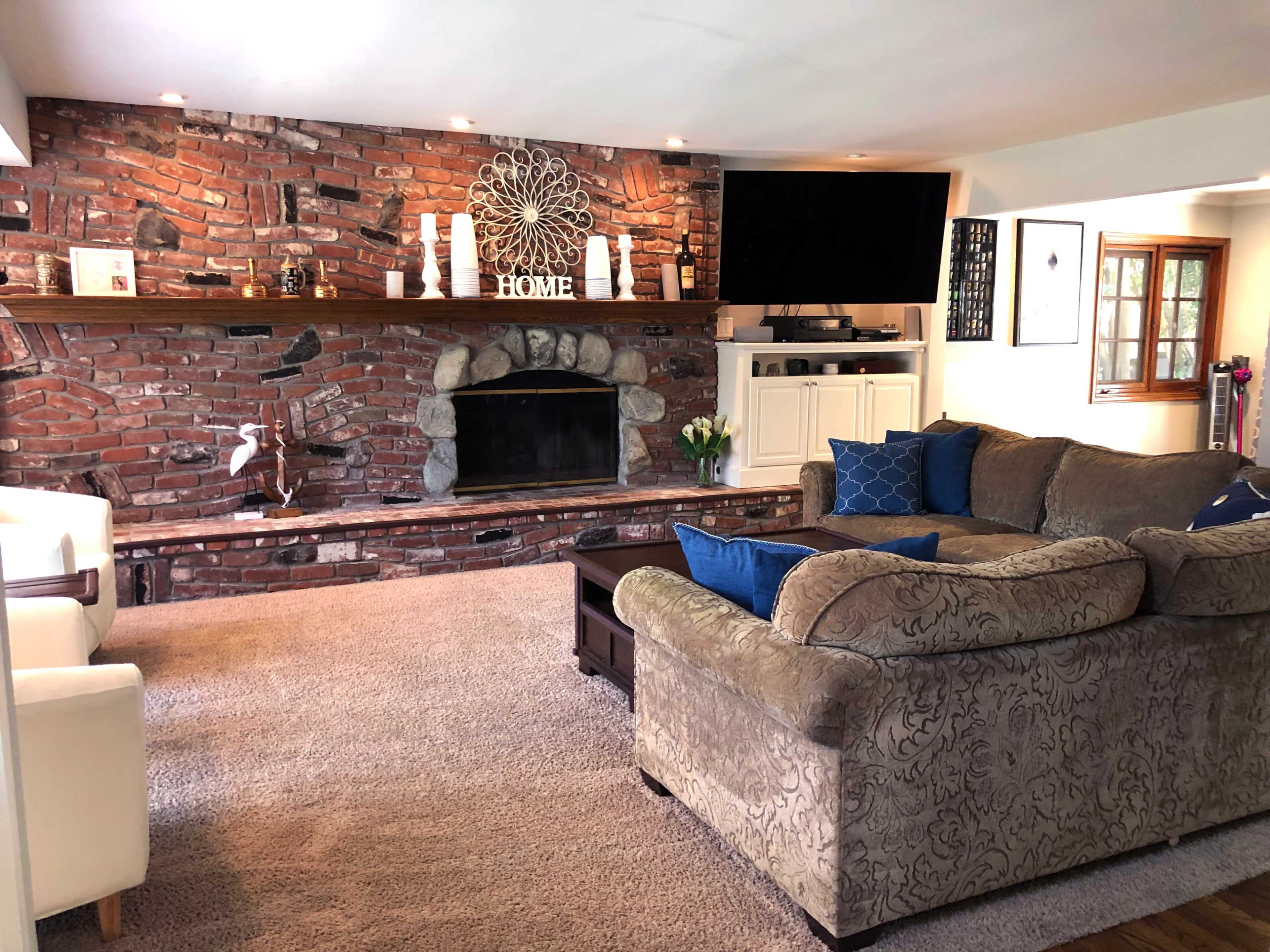 The image shows a cozy living room with a brick accent wall, a television mounted above a wooden shelf, and a comfortable sofa facing a stone fireplace.