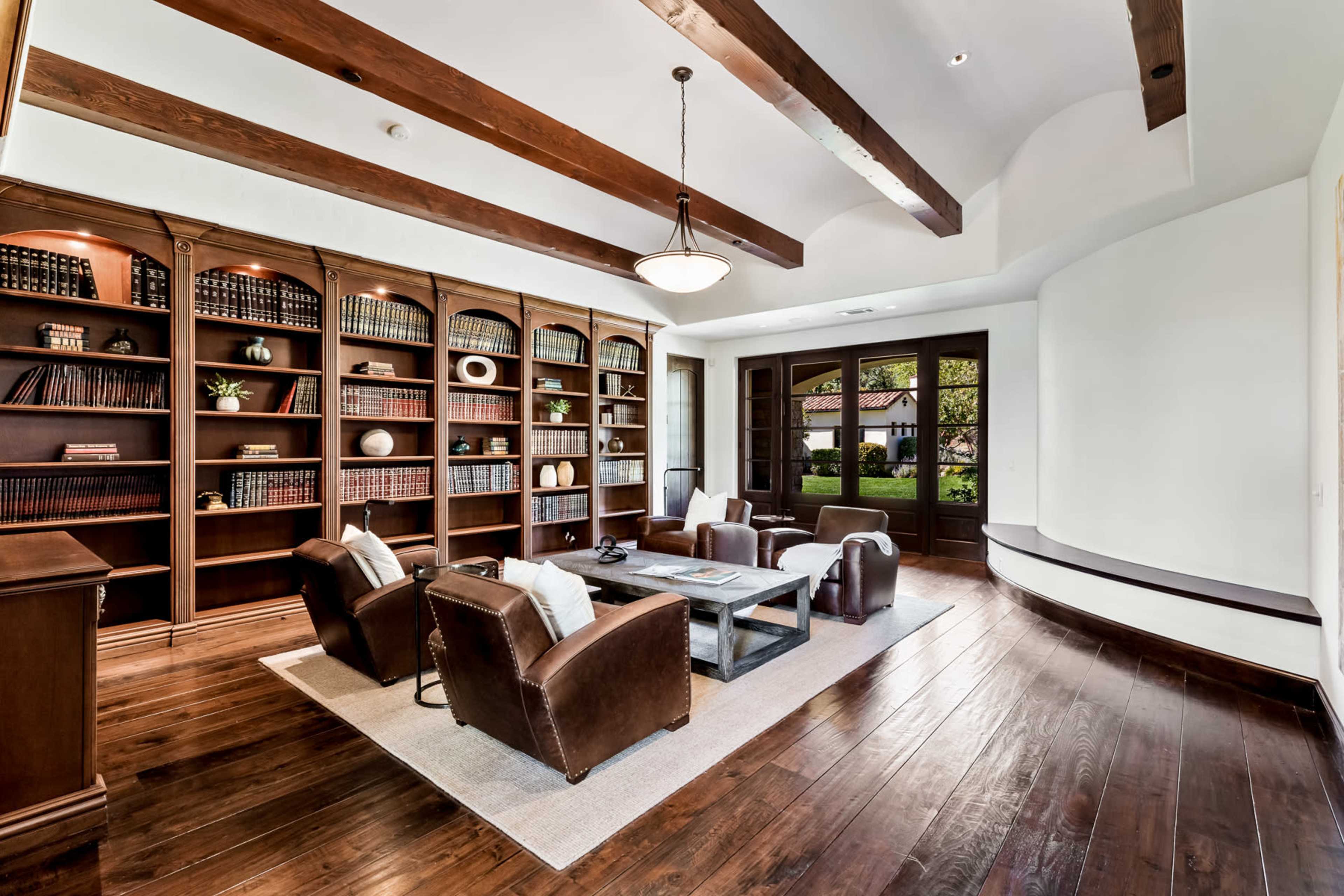 A room features wooden beams, a built-in bookshelf filled with books, and a seating area with leather chairs arranged around a coffee table on a rug.