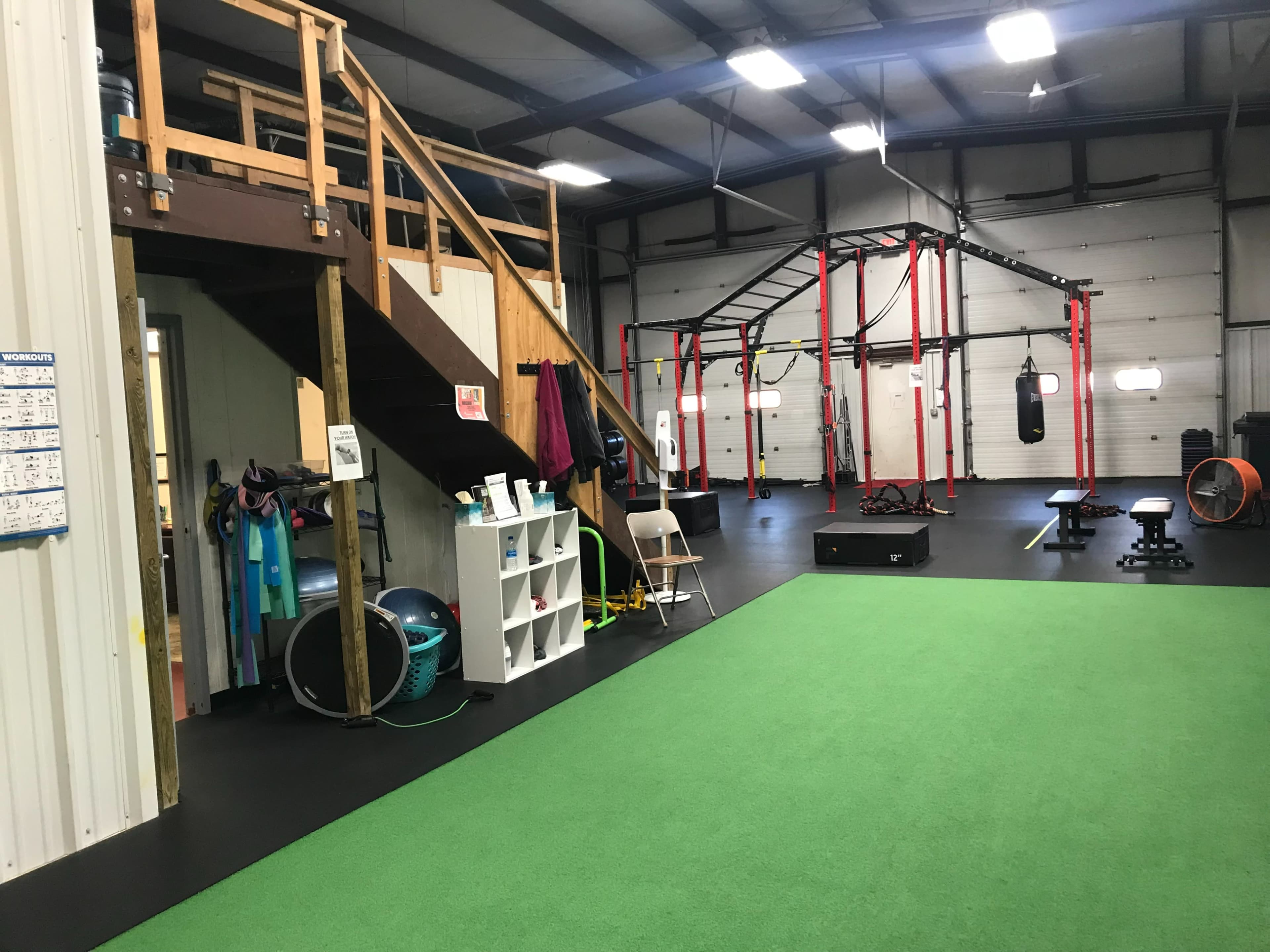 An indoor gym area featuring a turf floor, exercise equipment, a staircase leading to a loft, and various fitness apparatuses suspended from a frame.