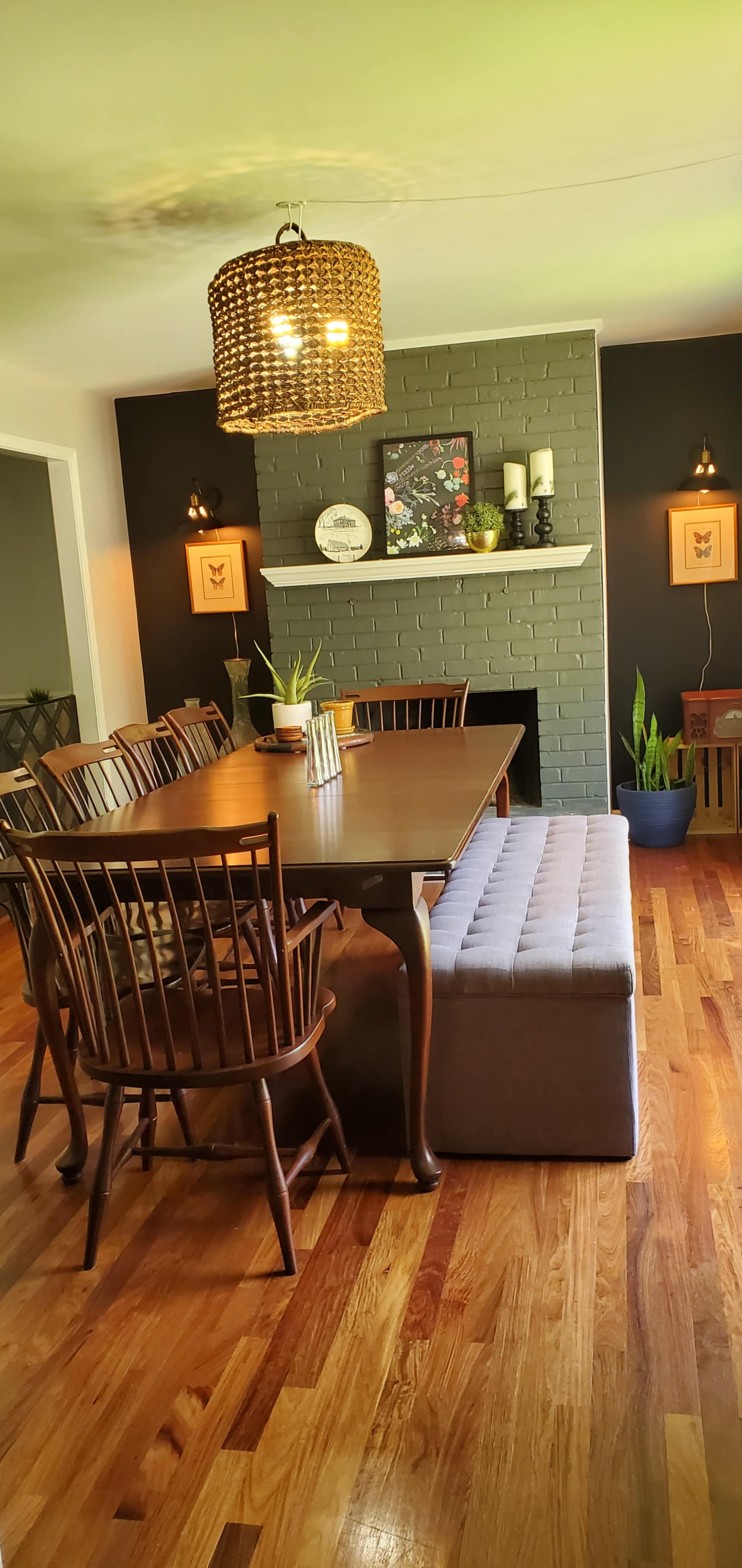 A wooden dining table with eight chairs is positioned in a room featuring a green accent wall with a fireplace and decorative plants.