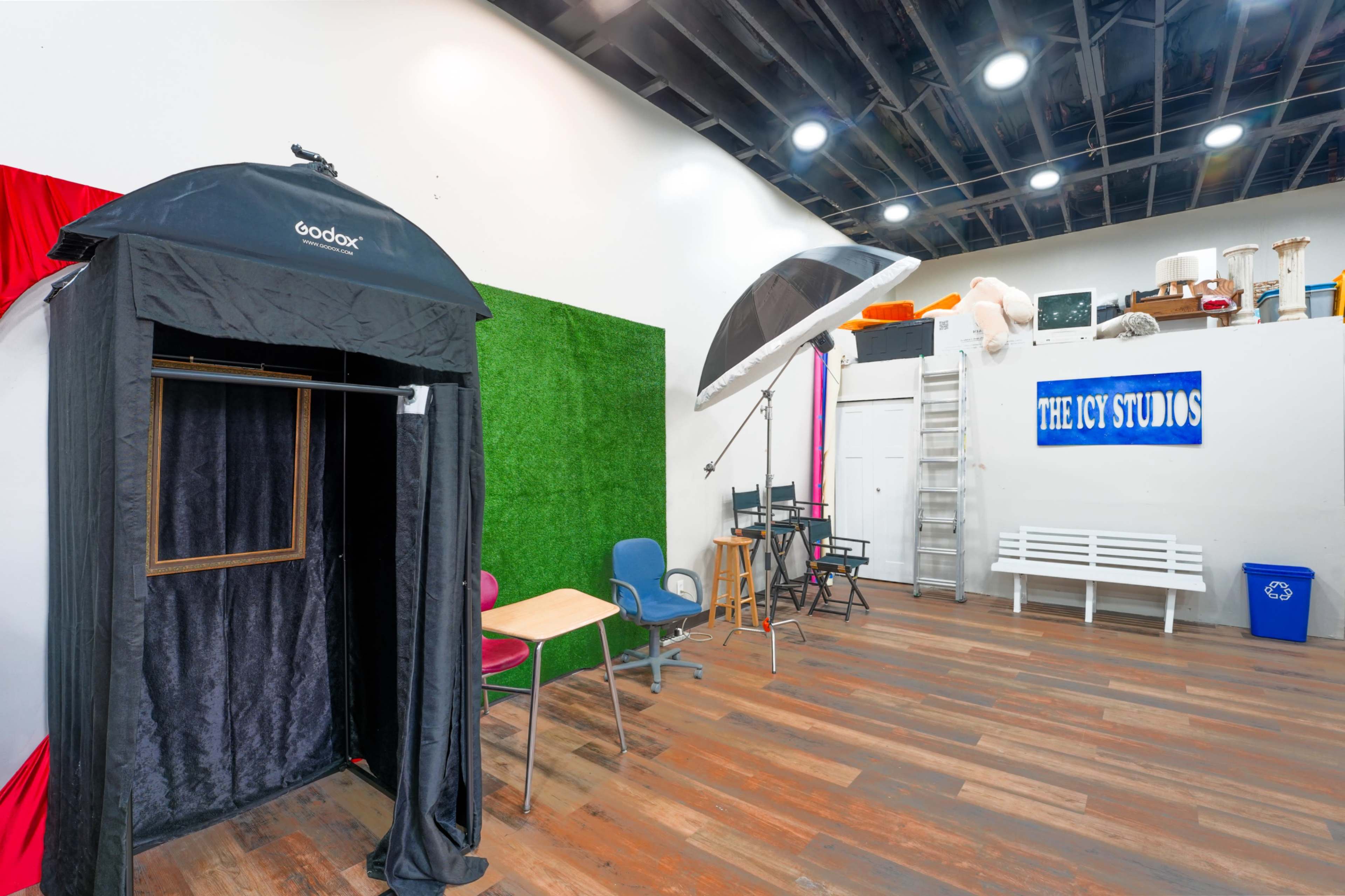 The image shows a studio space with a black photo booth, a large umbrella light, colorful chairs, and a decorative green wall.