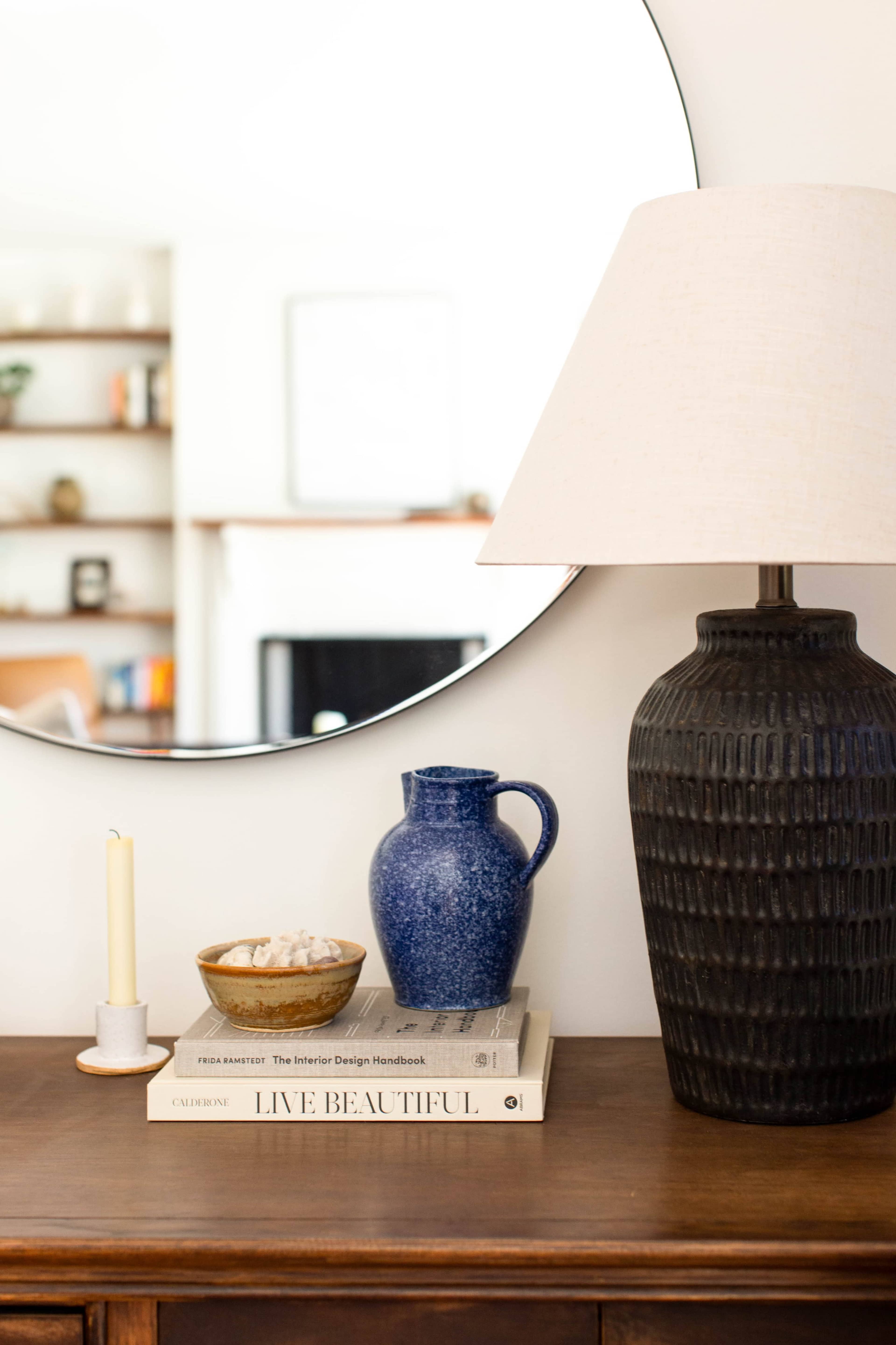 A wooden table holds a stack of design books, a lamp, a candle, a bowl, and a blue pitcher, with a round mirror reflecting a living room in the background.