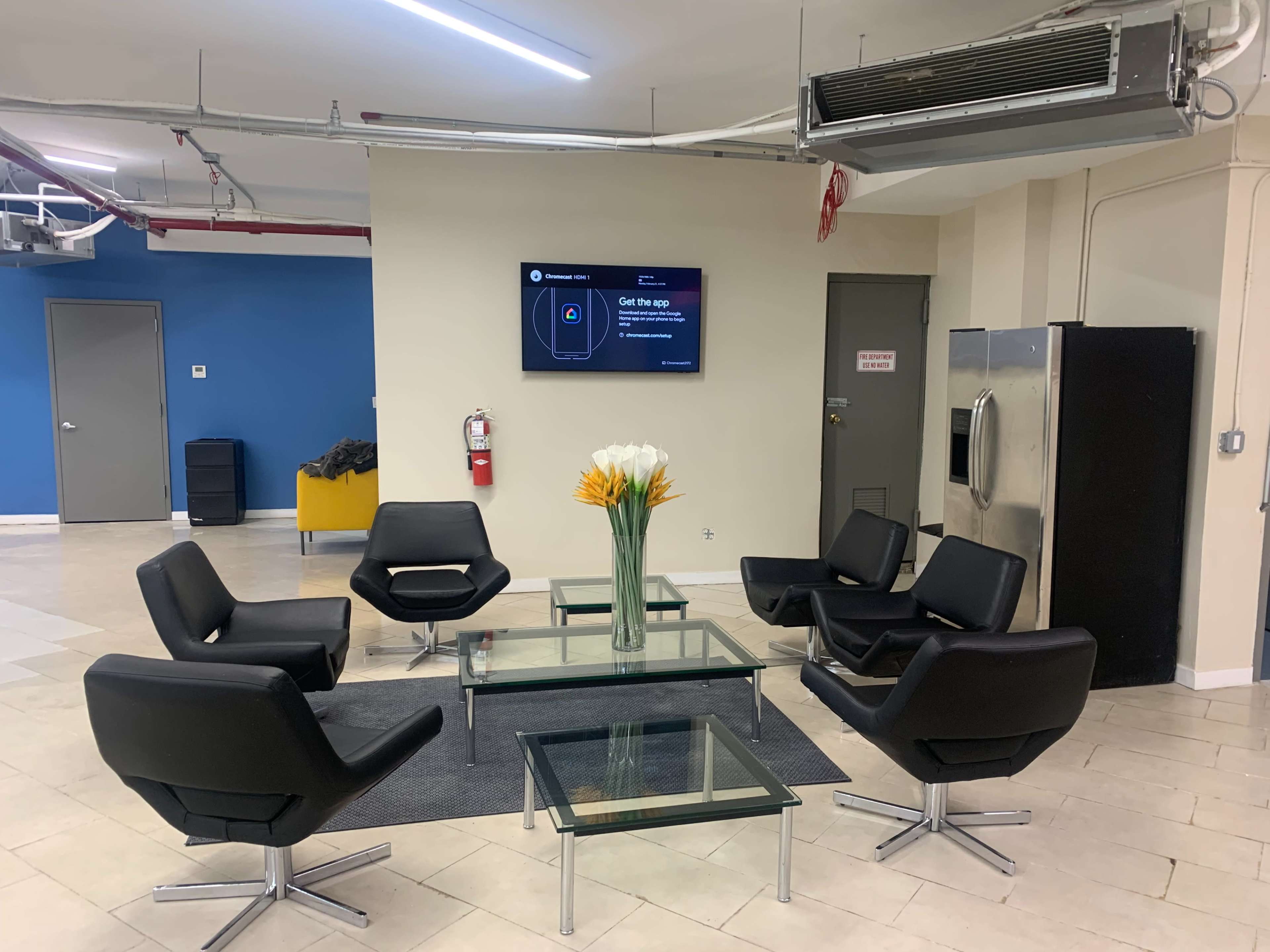 The image shows a waiting area with six black chairs arranged around a glass table, featuring a vase of yellow flowers, a wall-mounted screen, and a refrigerator.