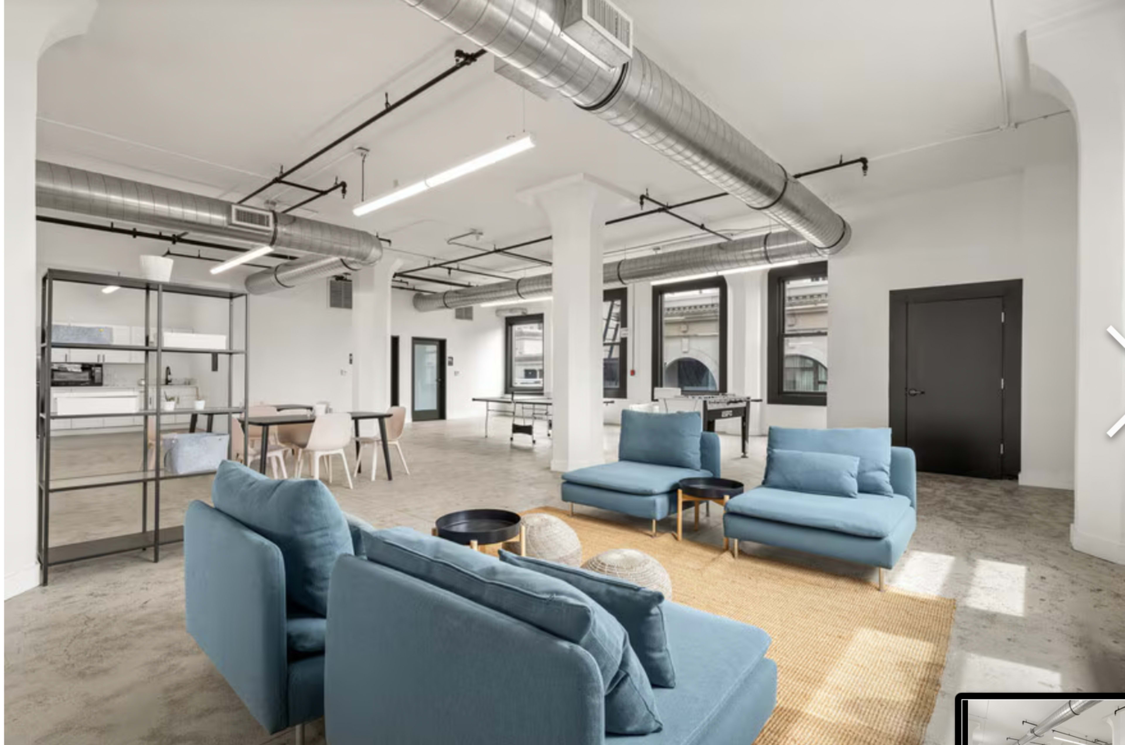 The image shows a spacious, modern interior with light-colored walls, industrial-style ventilation, and a seating area featuring blue sofas on a natural fiber rug.