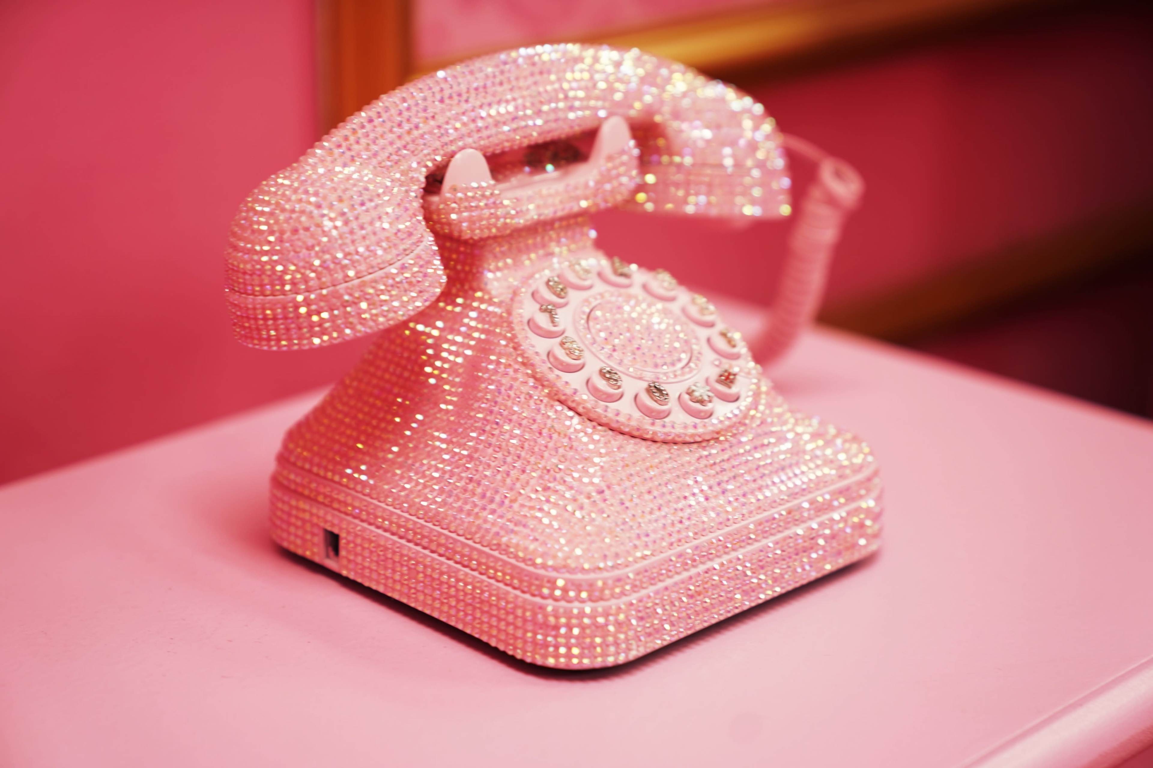A vintage rotary phone is covered in pink rhinestones and sits on a pink surface.