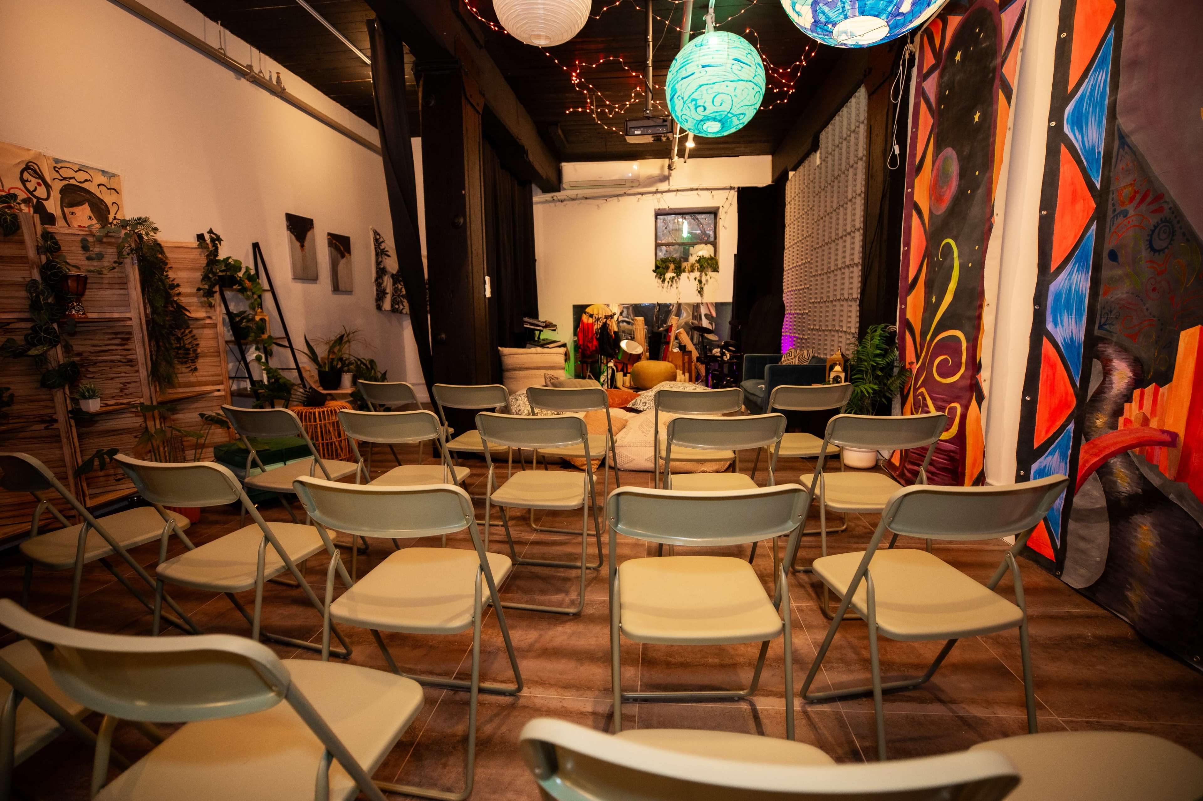 The image shows a colorful indoor space set up for an event, featuring rows of empty folding chairs facing a stage area adorned with plants and artwork.