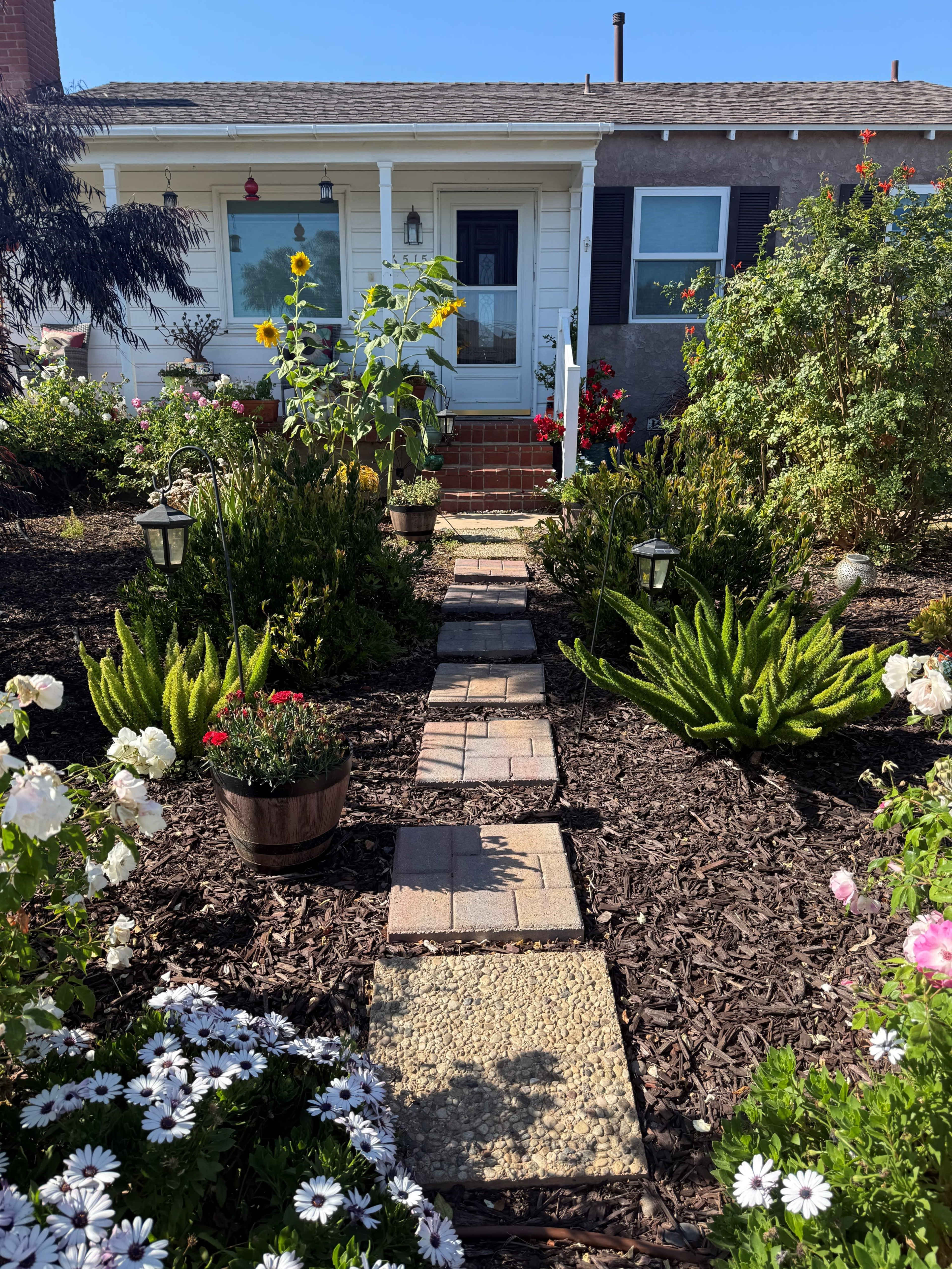 A stone pathway leads to a front porch, flanked by various flower beds and plants in a garden setting.