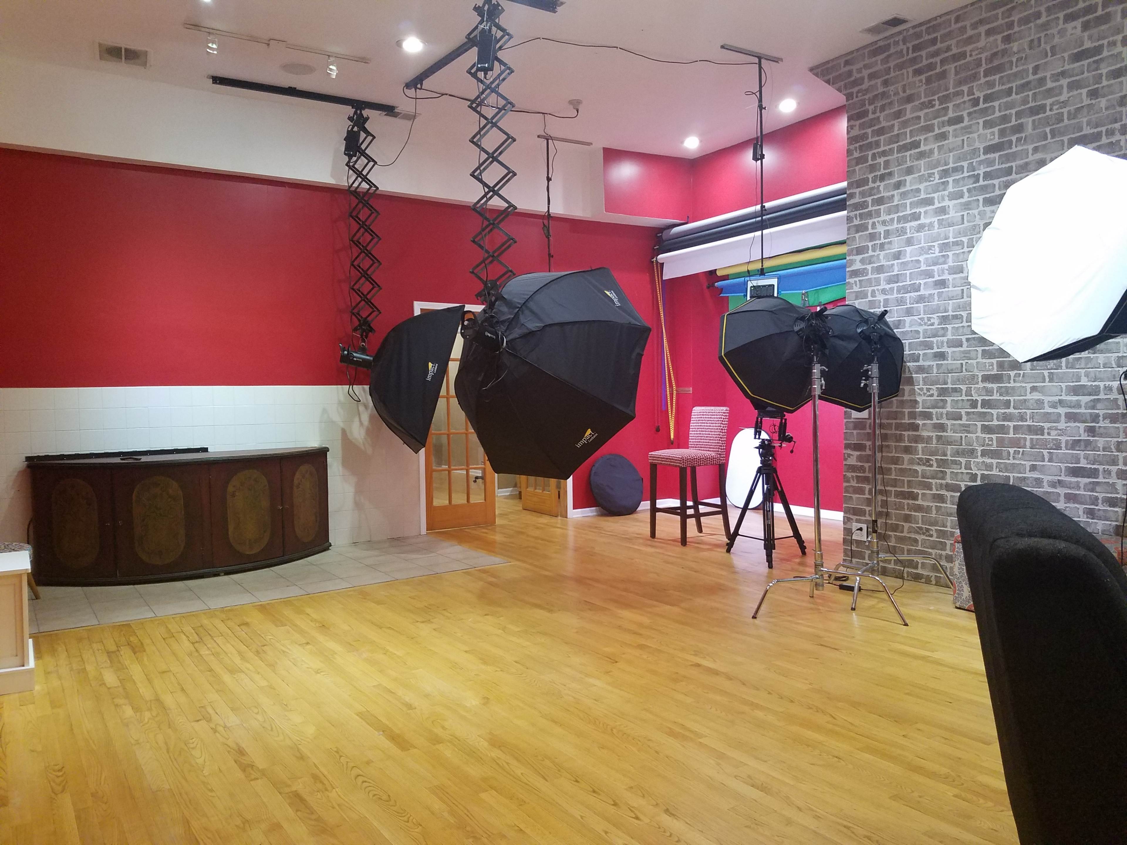 The scene features a photography studio with large softbox lights, a brick wall, and wooden flooring, alongside various furniture pieces.