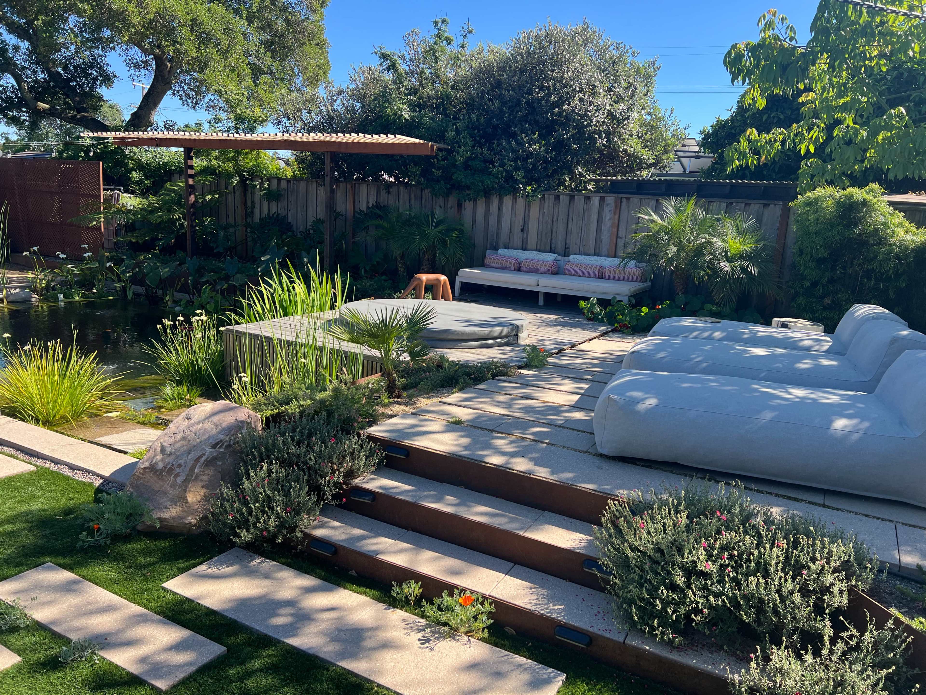 The image shows a landscaped backyard featuring a pond, seating areas, and various plants arranged around a stone pathway.
