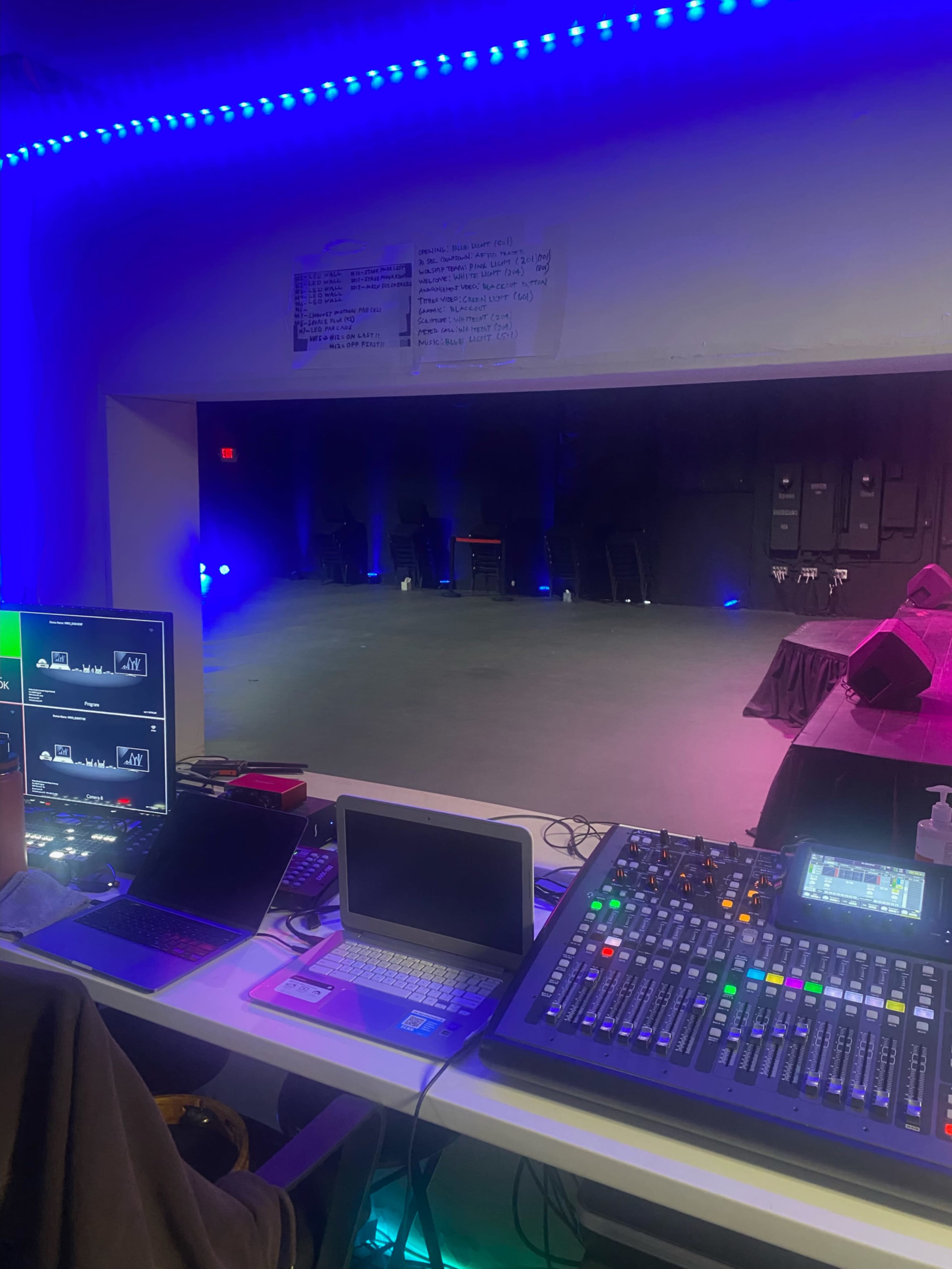 The image shows a sound booth with various equipment, including monitors and a mixing console, overlooking a dimly lit stage area with colored lighting.