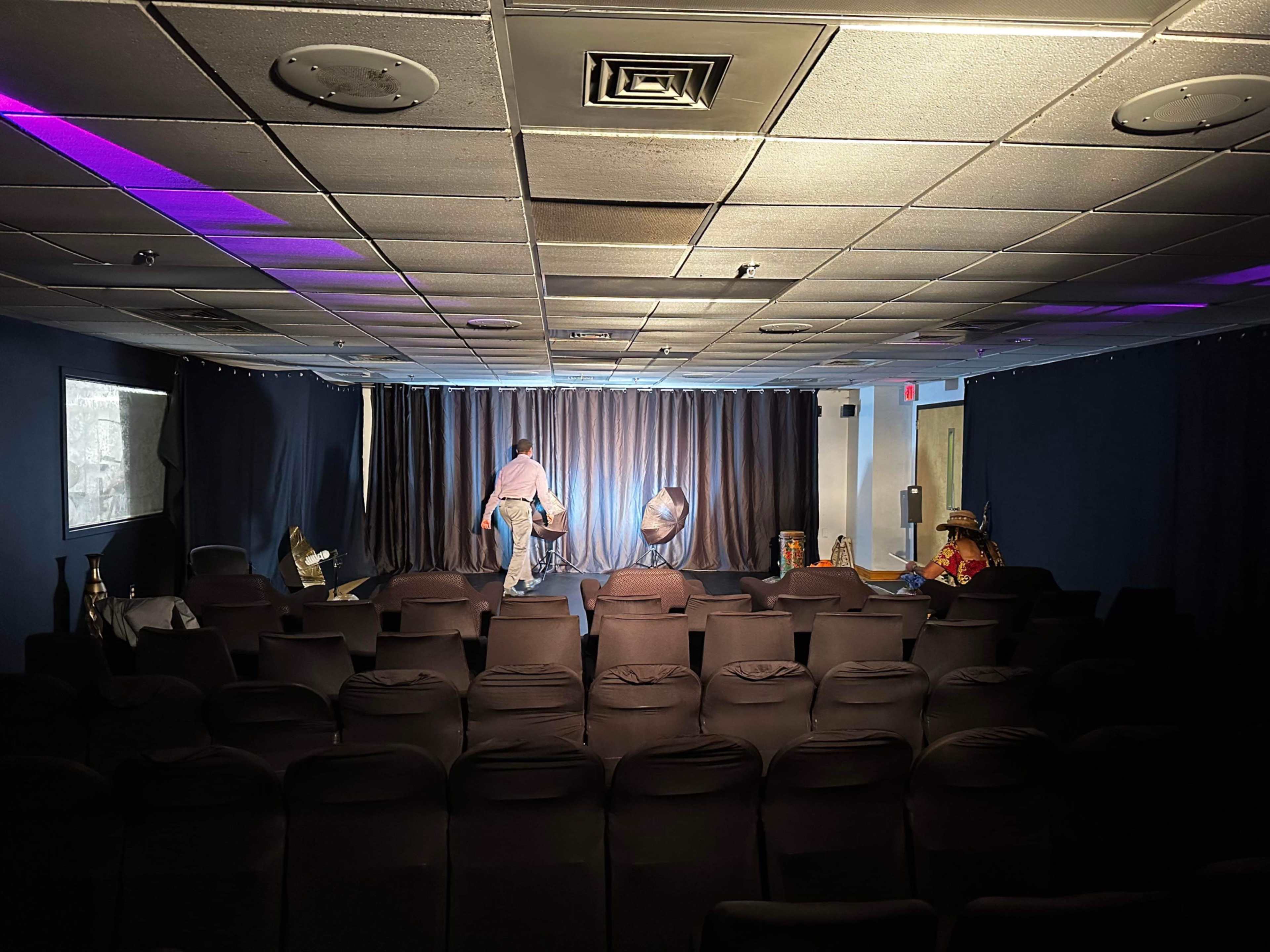 A small theater space with black chairs, a stage draped in curtains, and a performer setting up props under dim lighting.