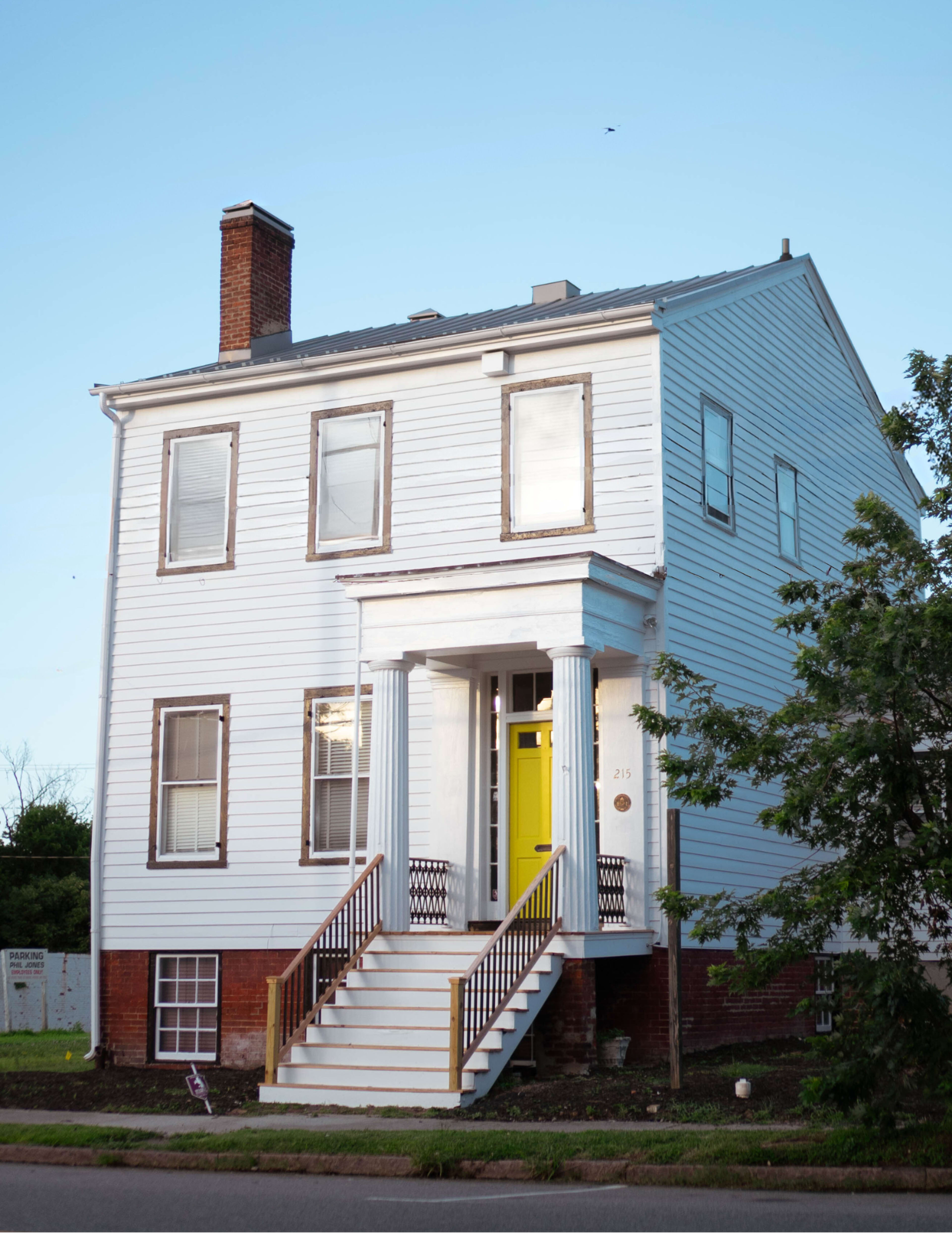 A two-story white house with a yellow front door and a set of stairs leads up to a porch, surrounded by green shrubs.