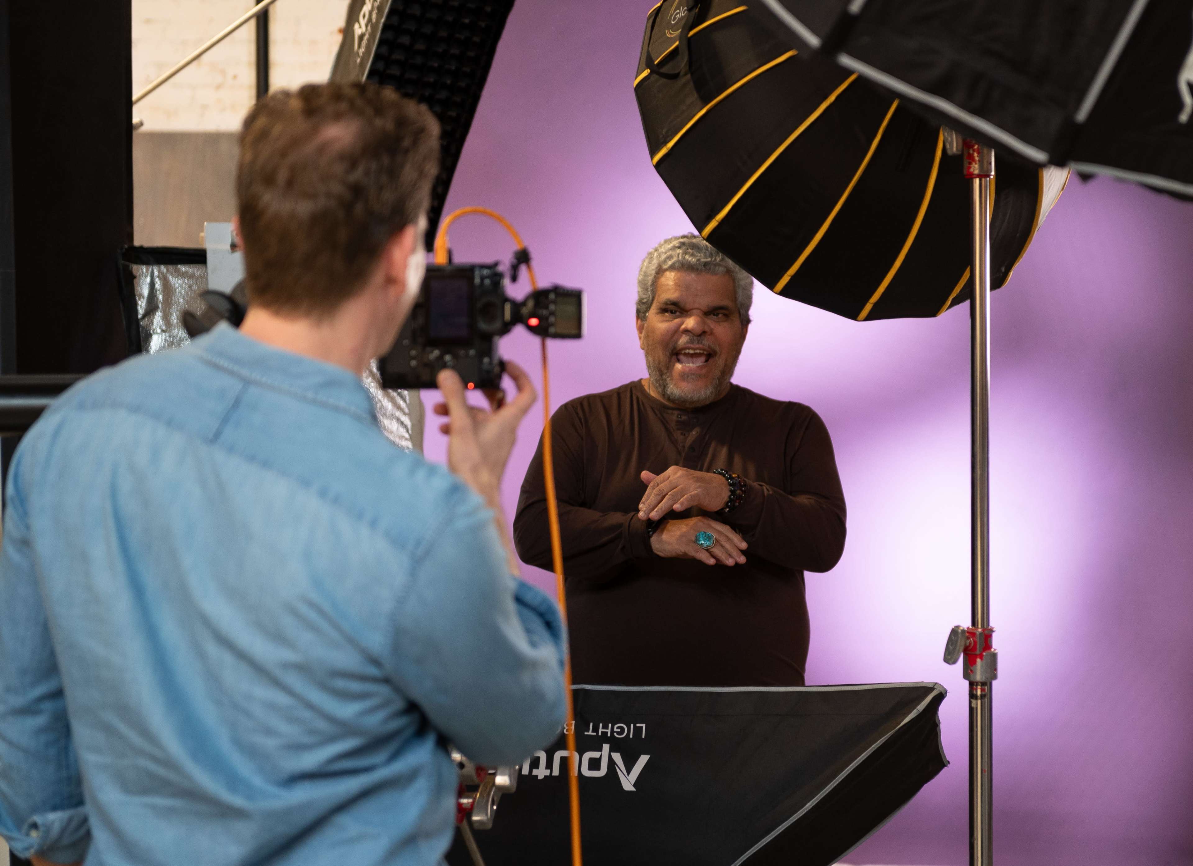 A photographer is capturing a portrait of a smiling man in front of a soft purple backdrop, with studio lighting equipment positioned around them.