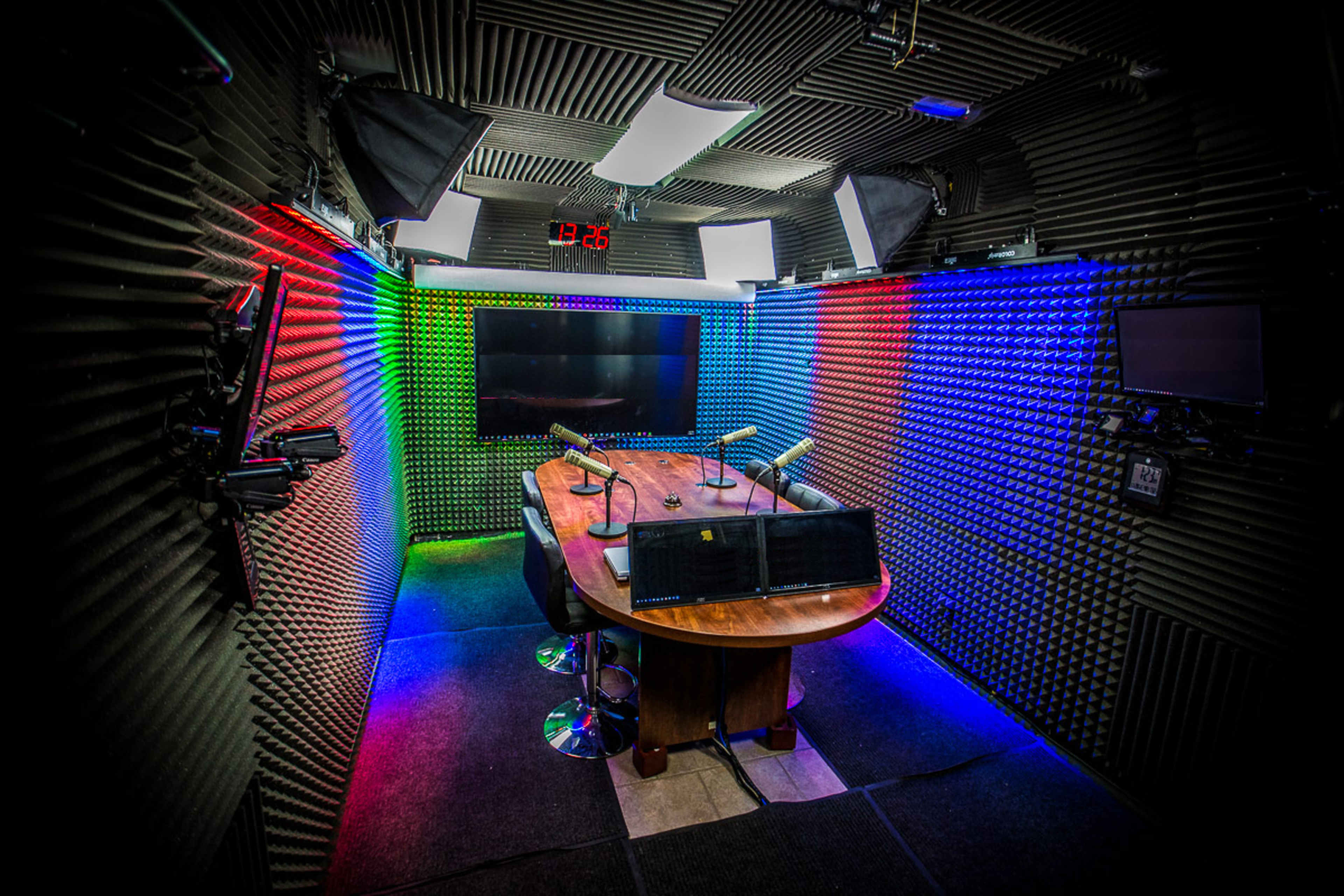The image shows a modern recording studio with soundproof walls, a central wooden table, and various lighting fixtures illuminating the space.