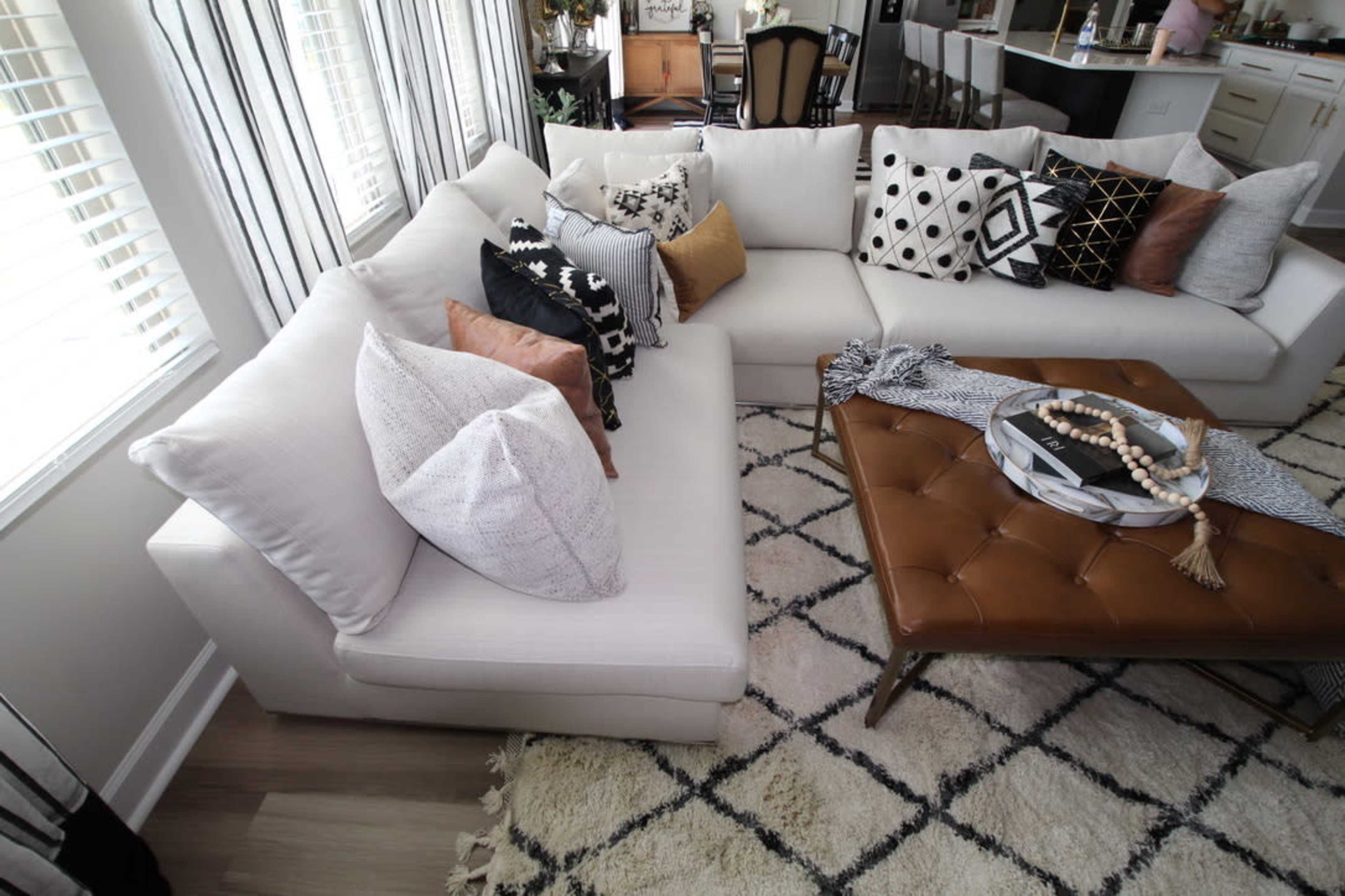 A modern living room featuring a light-colored sectional sofa adorned with various patterned and textured cushions, paired with a dark, tufted coffee table.