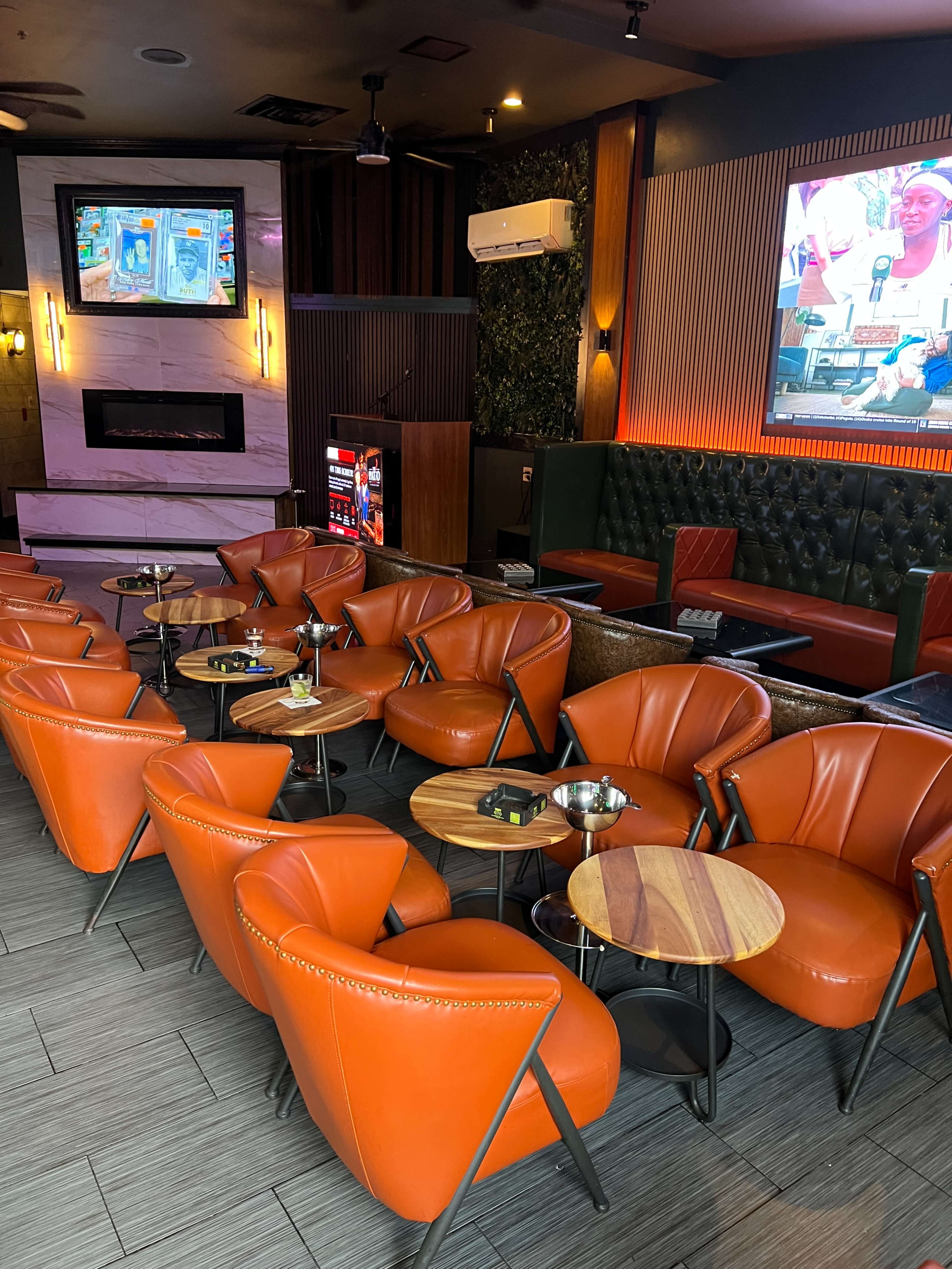 The image shows a stylish lounge area featuring orange chairs arranged around wooden tables, with a fireplace and screens displaying content in the background.