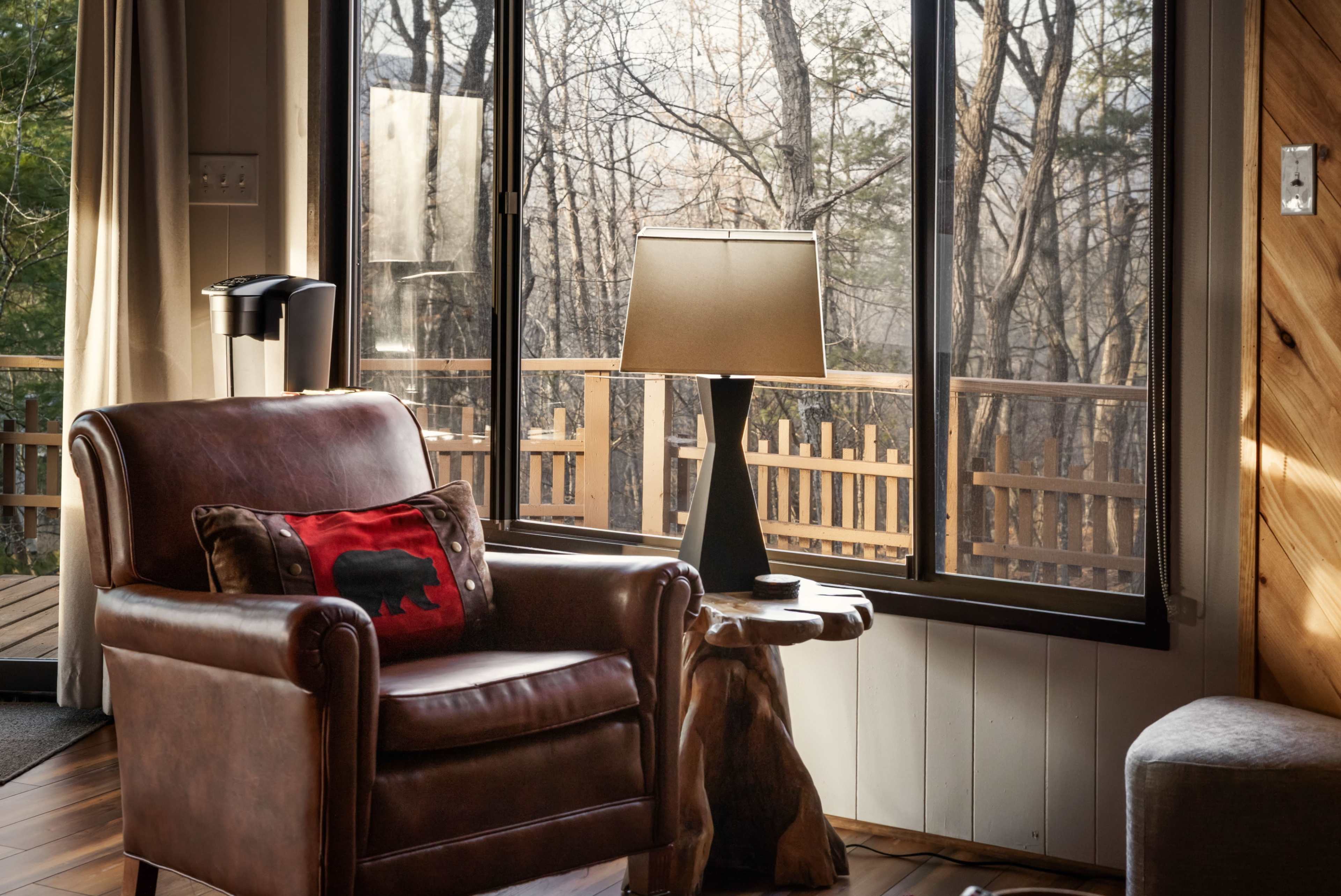 A leather armchair with a decorative pillow sits beside a wooden side table next to a window overlooking a forested area.