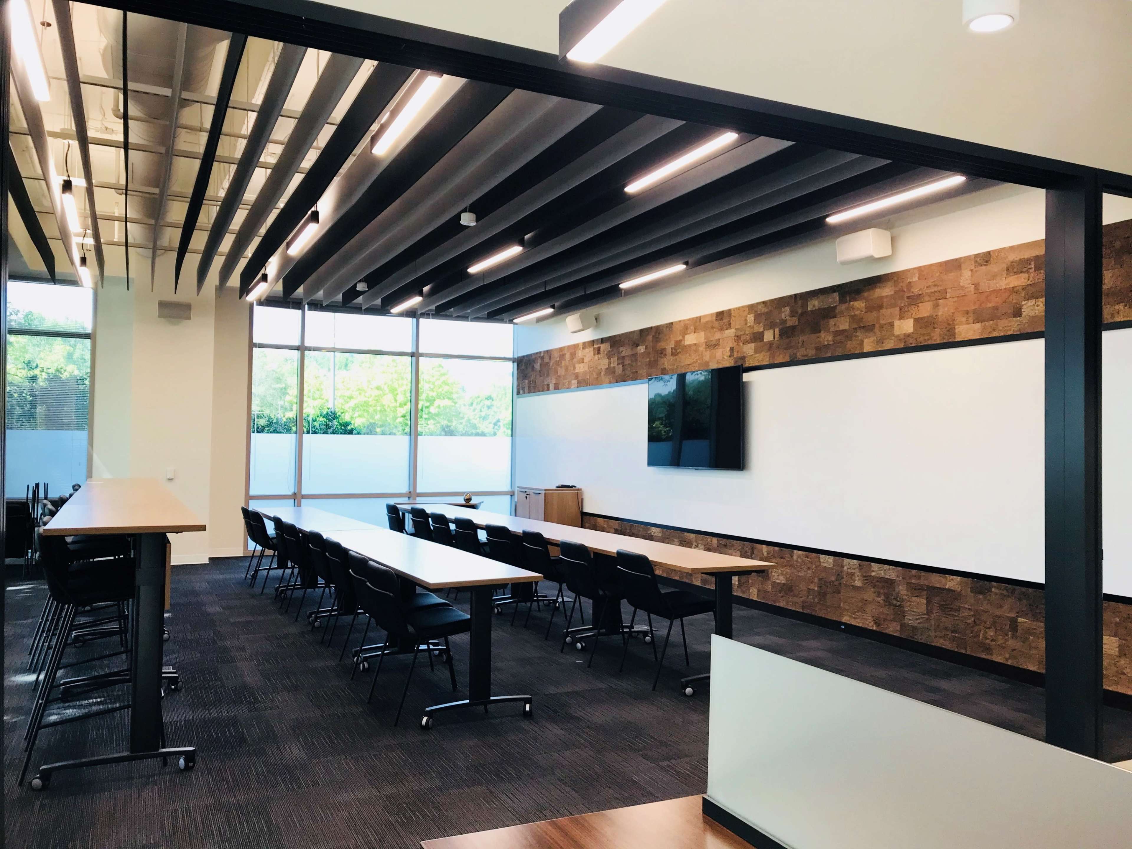 A large classroom features multiple tables and chairs arranged for seating, with a presentation screen and windows allowing natural light.