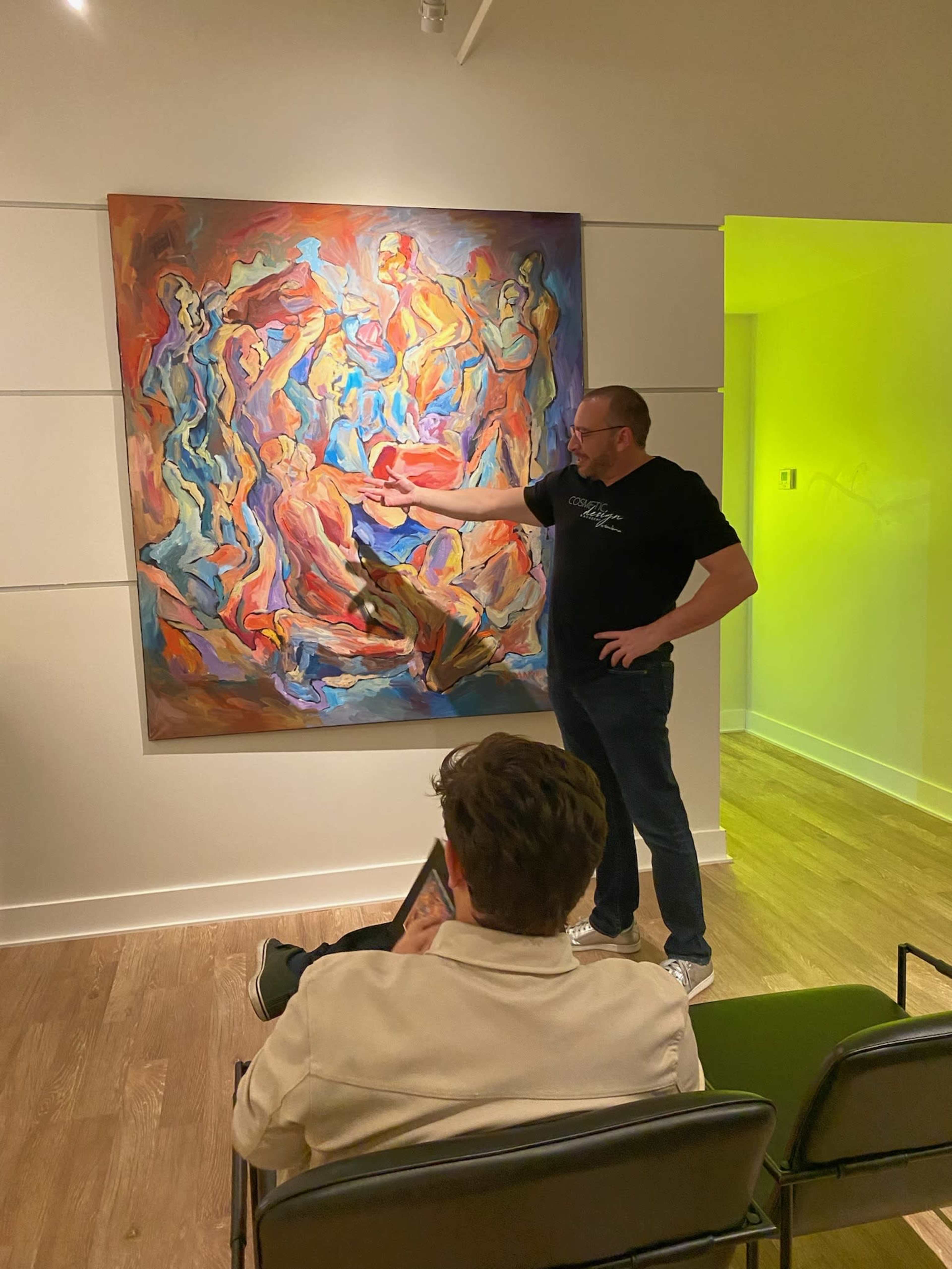 A man points at an abstract painting while another person sits on a chair holding a tablet.