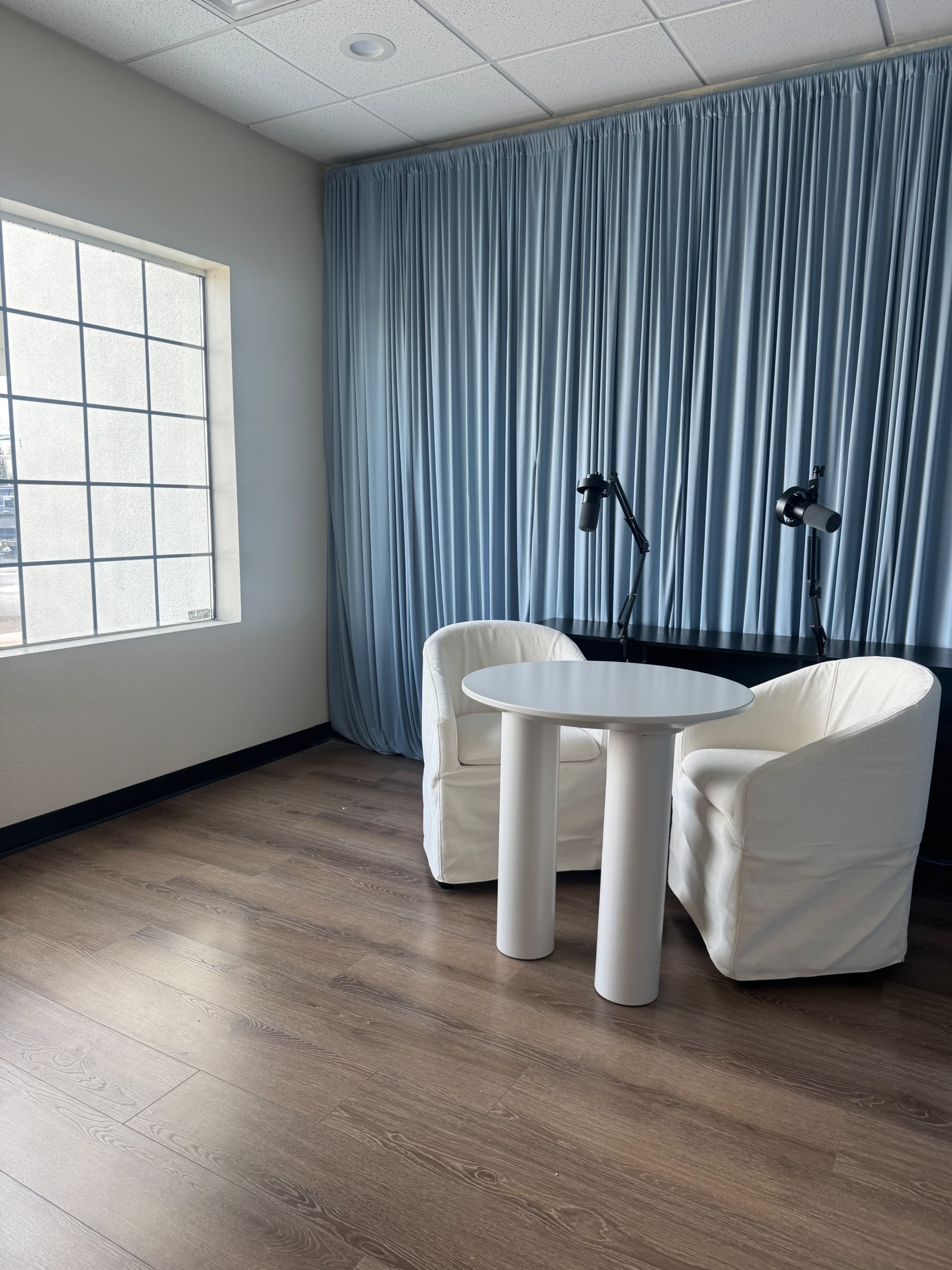 The image shows a small room with two white chairs and a round table, illuminated by natural light through a large window.