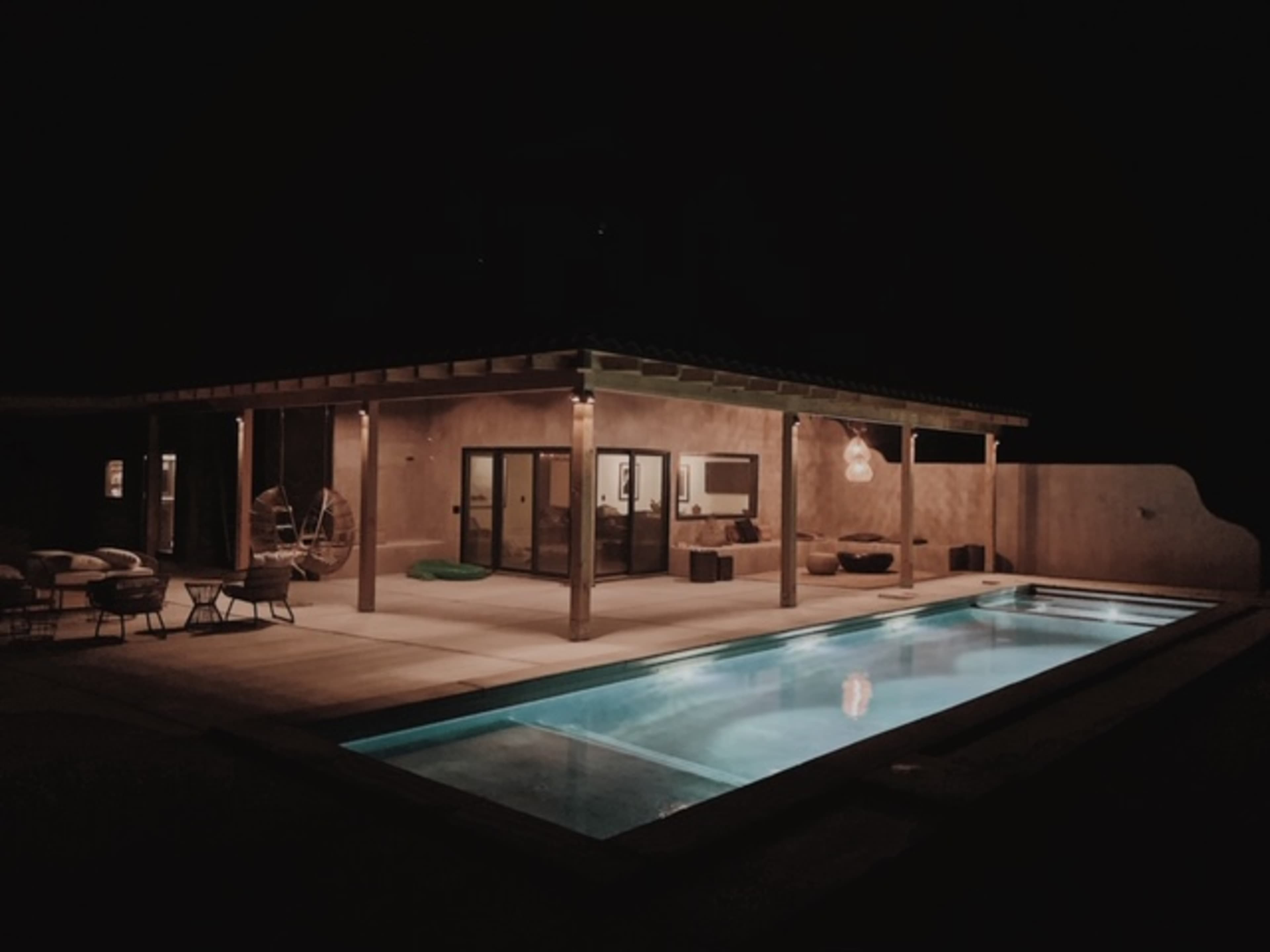 A modern outdoor space featuring a pool and a covered patio with furniture, illuminated softly at night.
