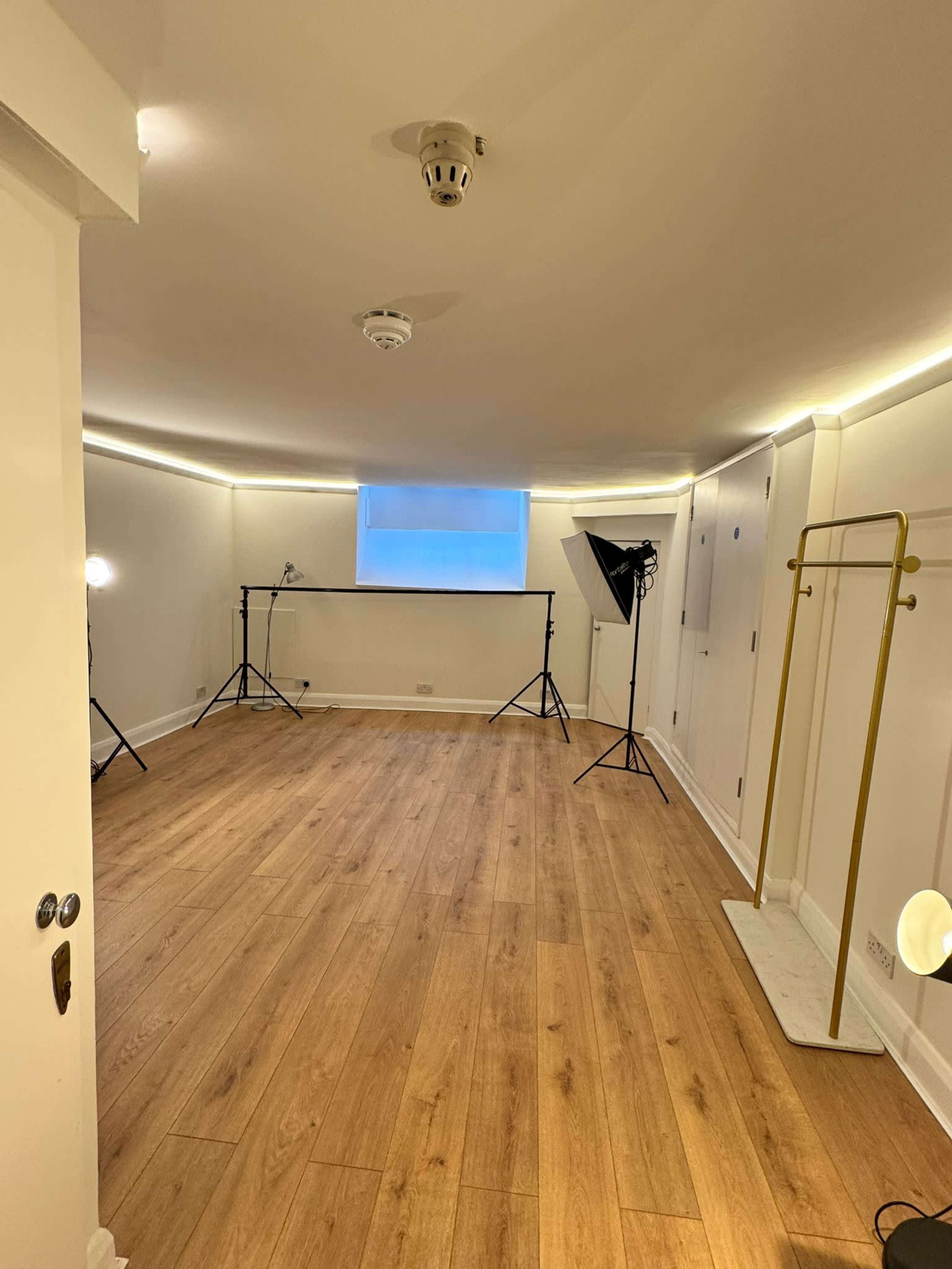 The image shows a blank, well-lit interior room featuring wooden flooring, two photography lights on stands, and a clothing rack against a white wall.
