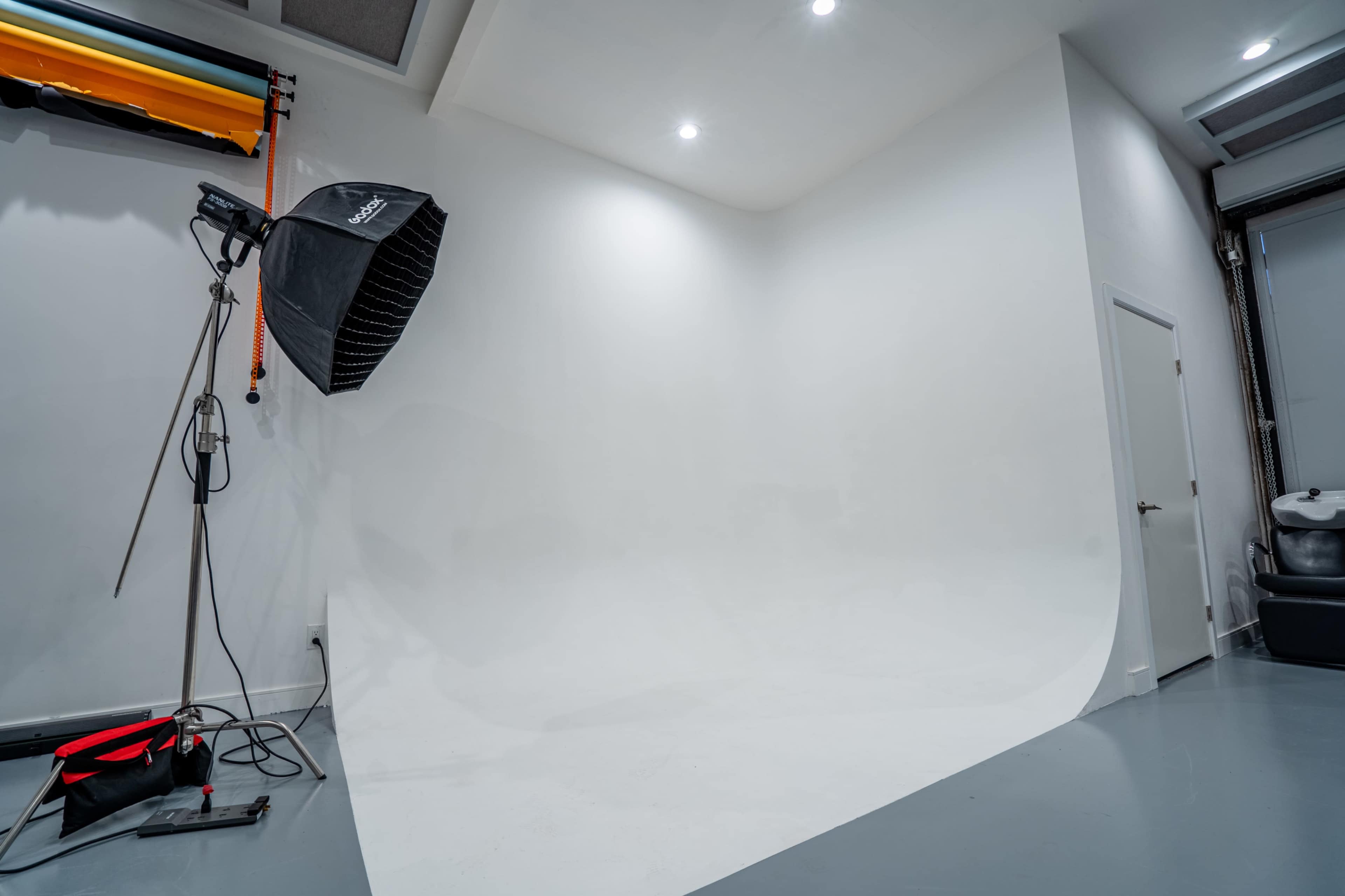 The image shows a photography studio with a seamless white backdrop and a lighting setup, including a softbox and a light stand.