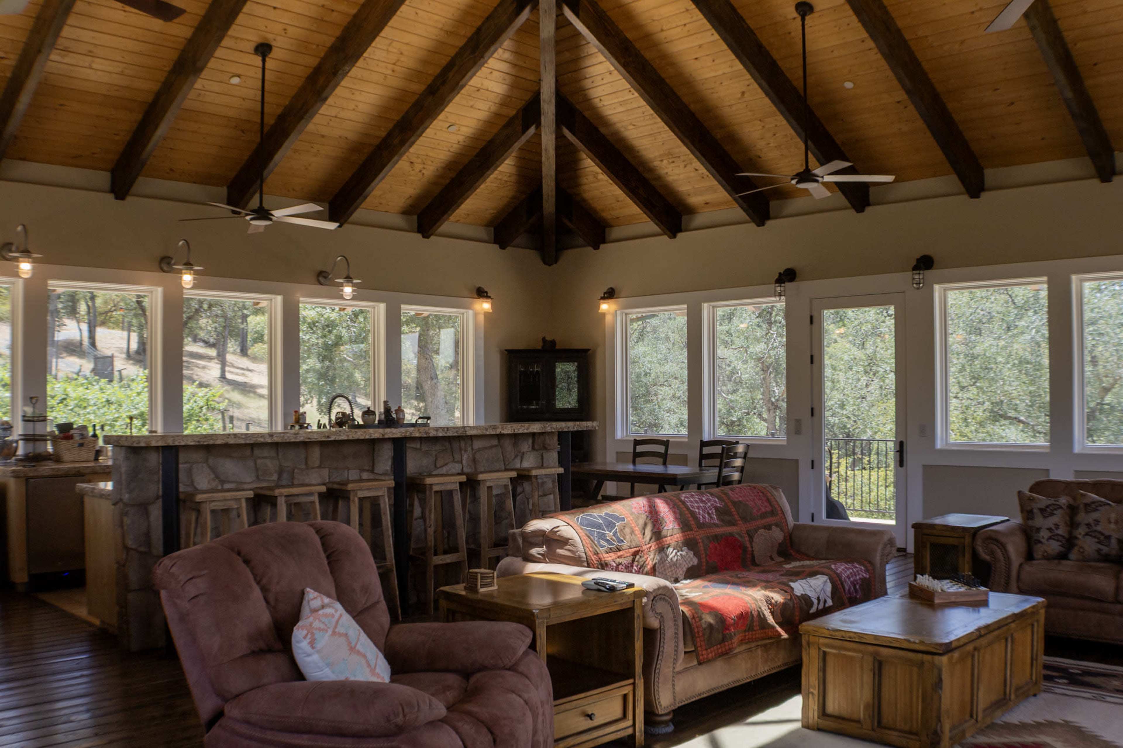 The image shows a spacious living area with a wooden ceiling, large windows, and a bar, featuring comfortable seating arrangements and a coffee table.