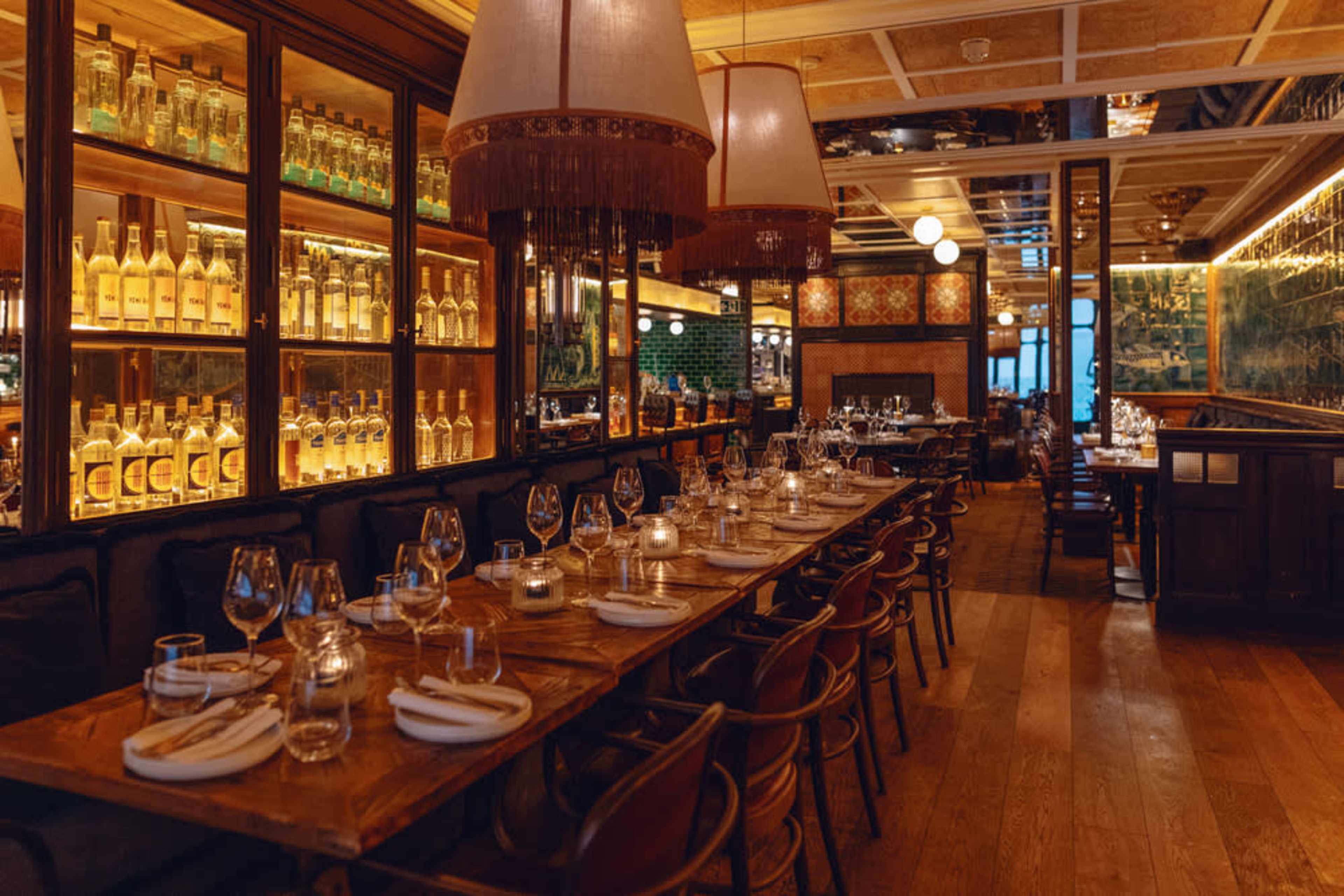 A warmly lit restaurant interior with a long wooden table set for dining, surrounded by shelves displaying bottles and stylish pendant lights.