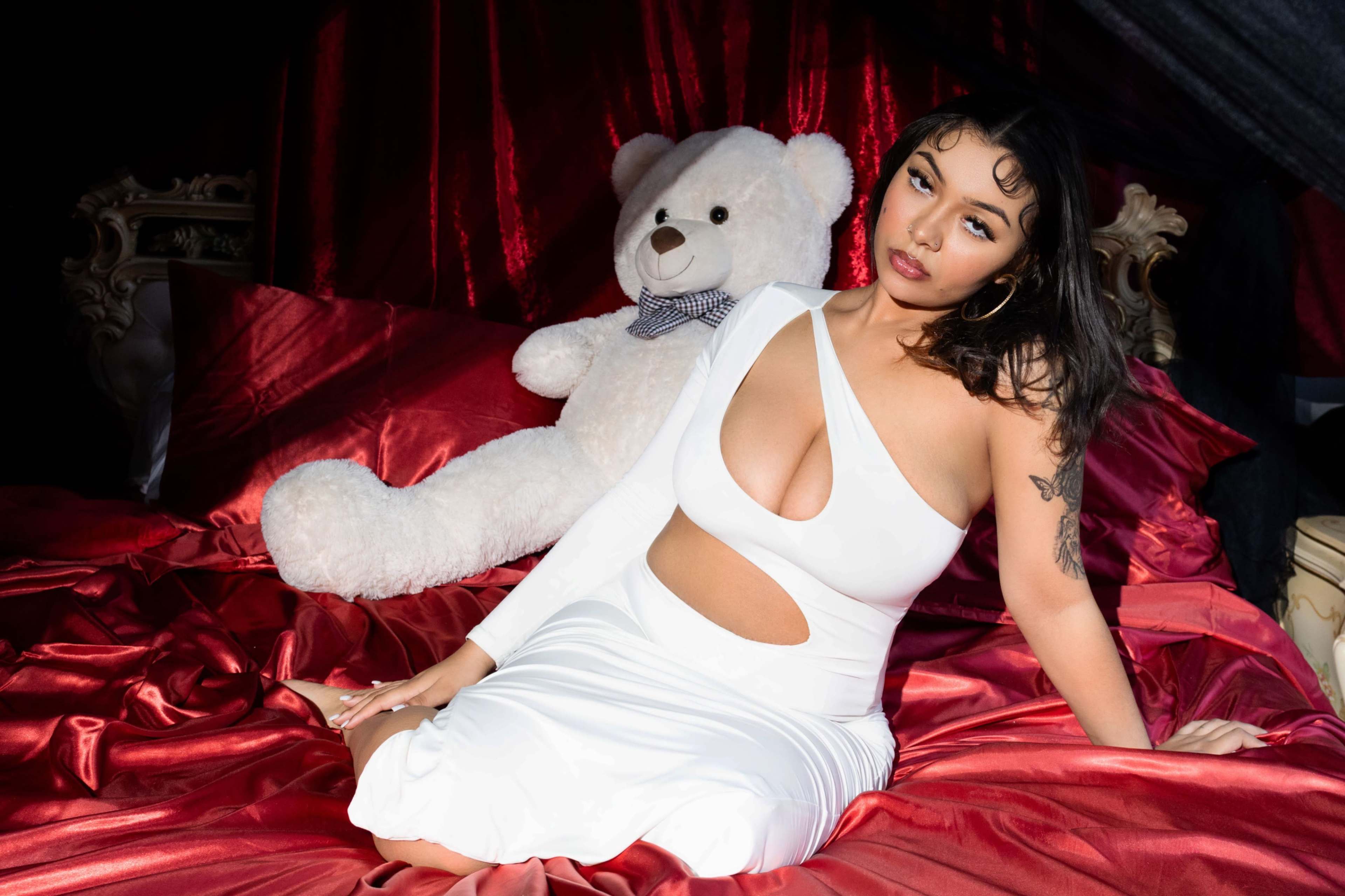A woman in a white dress sits on a red bed with a large stuffed bear beside her.