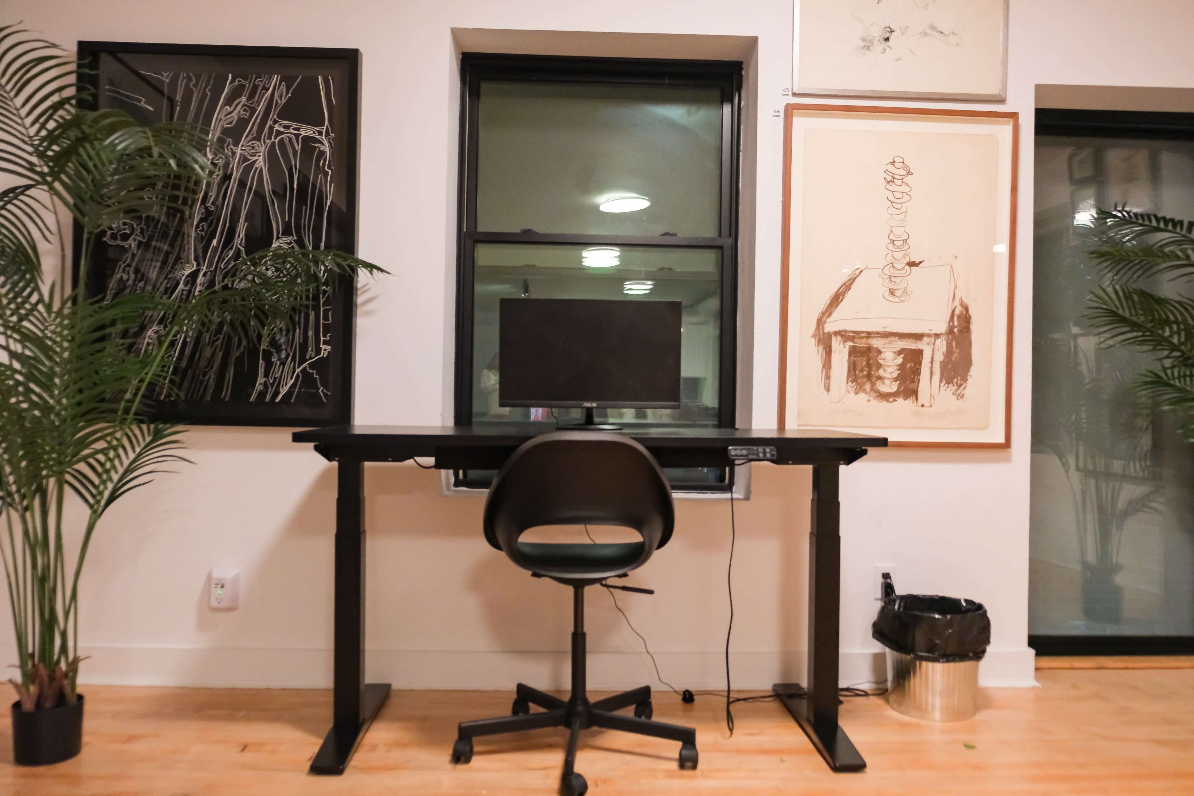 A black standing desk with a computer monitor and a rolling chair is positioned in front of a window, flanked by two framed artworks and potted plants.
