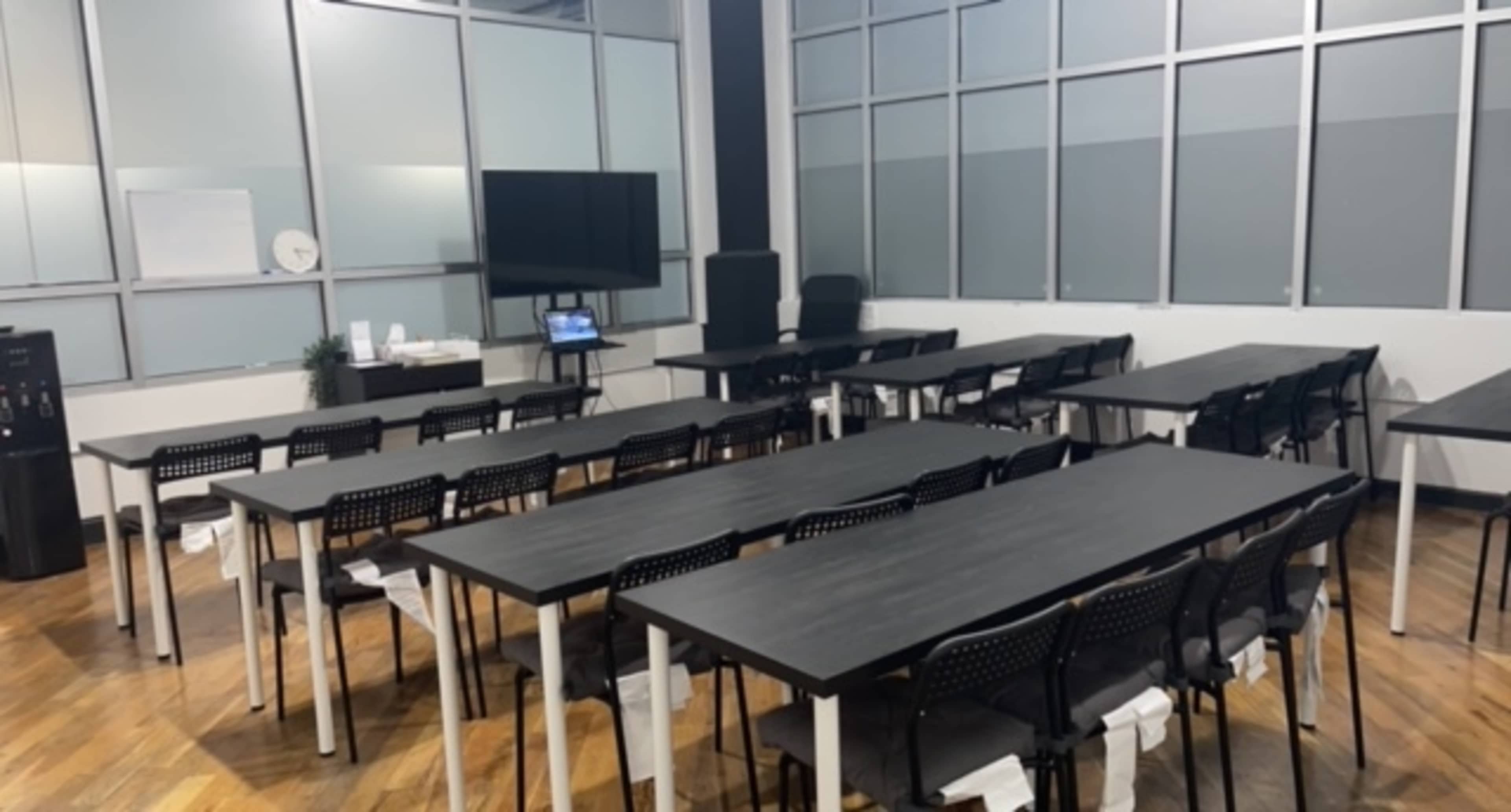 A large meeting room with multiple black tables and chairs arranged in a grid pattern, along with a television and water cooler at one end.