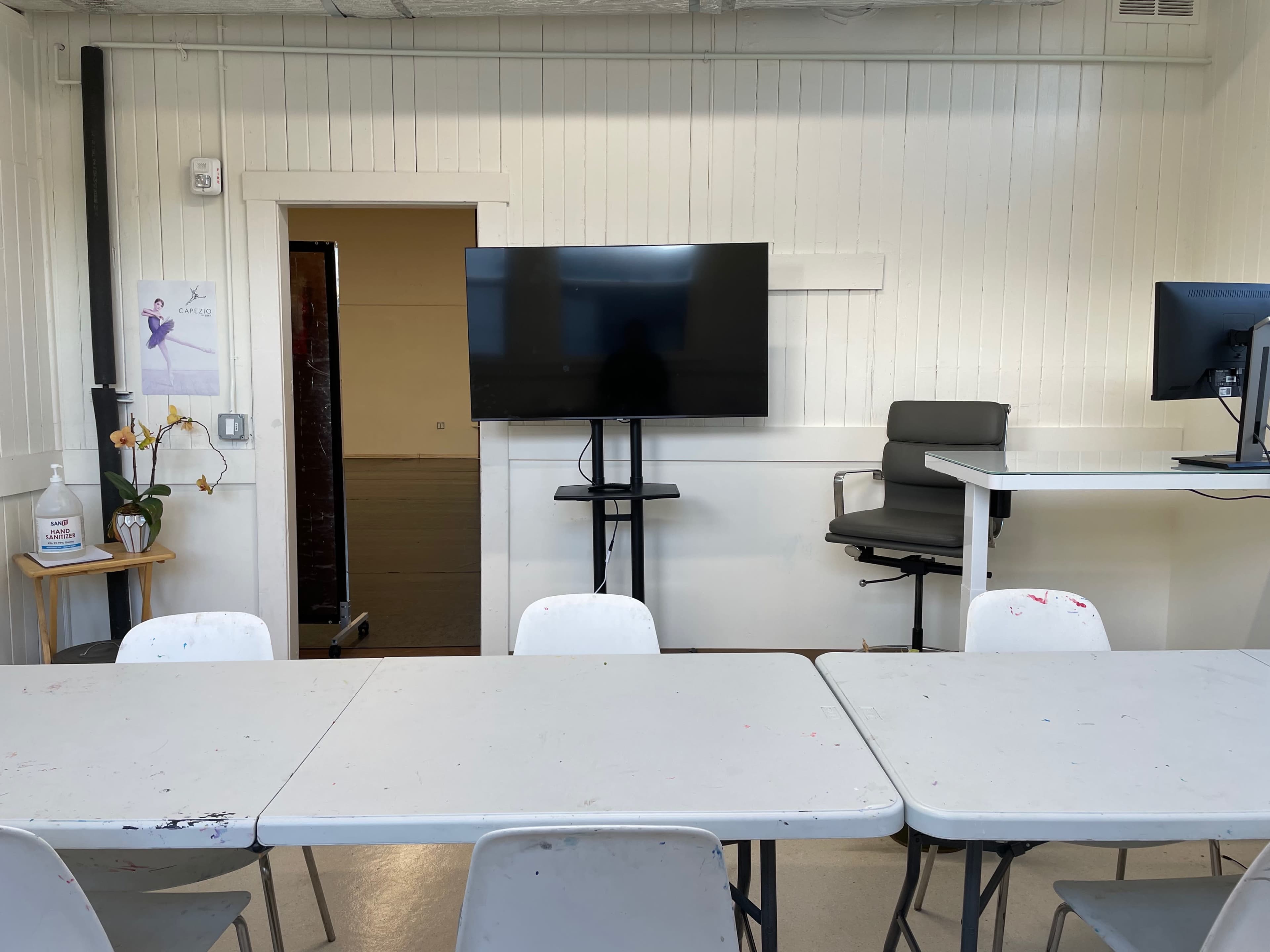 A clean, well-organized classroom with tables, chairs, a large TV screen, and a doorway leading to another room.