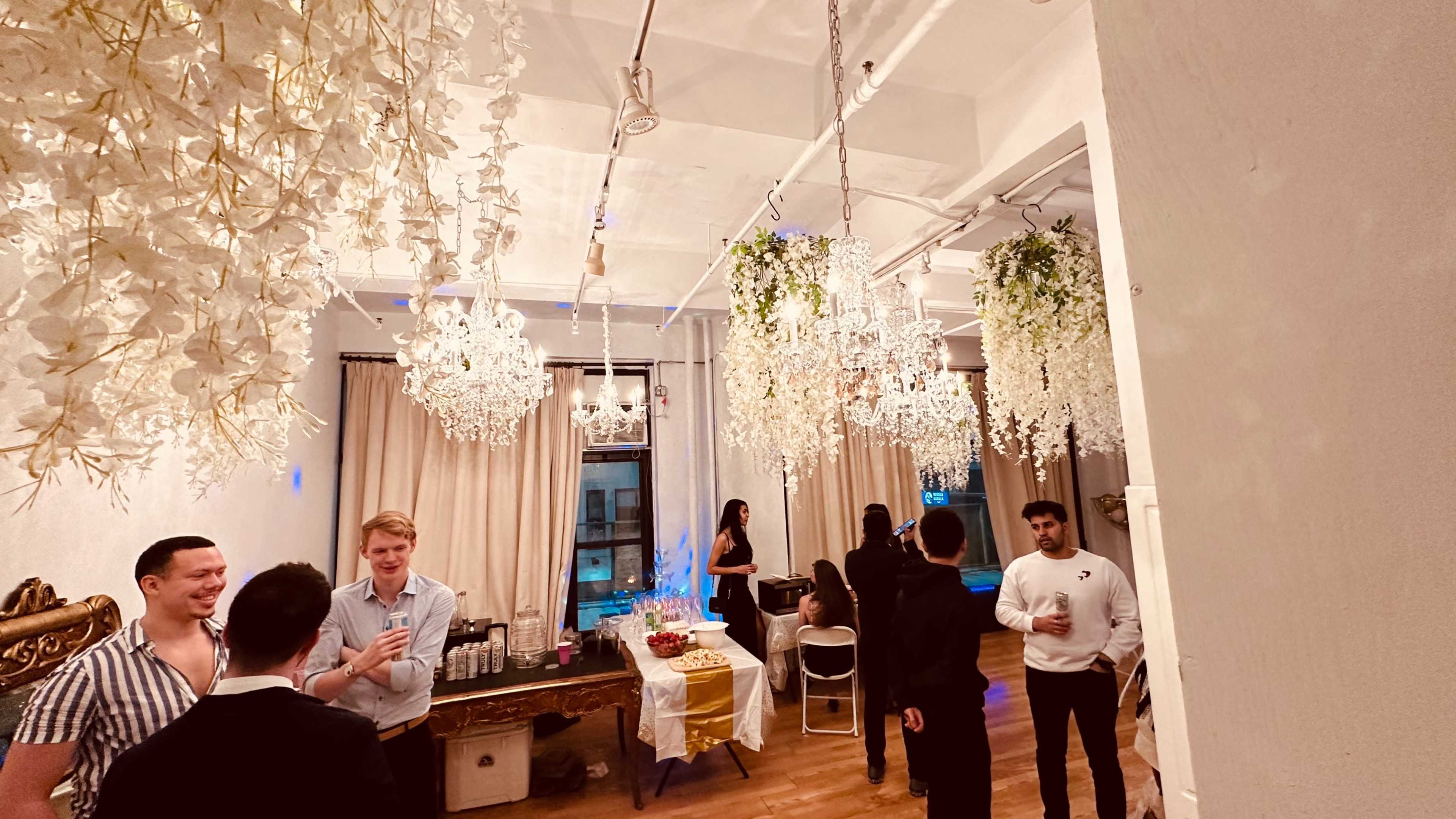 A gathering takes place in a decorated room with hanging floral arrangements and chandeliers, as guests converse and enjoy refreshments.
